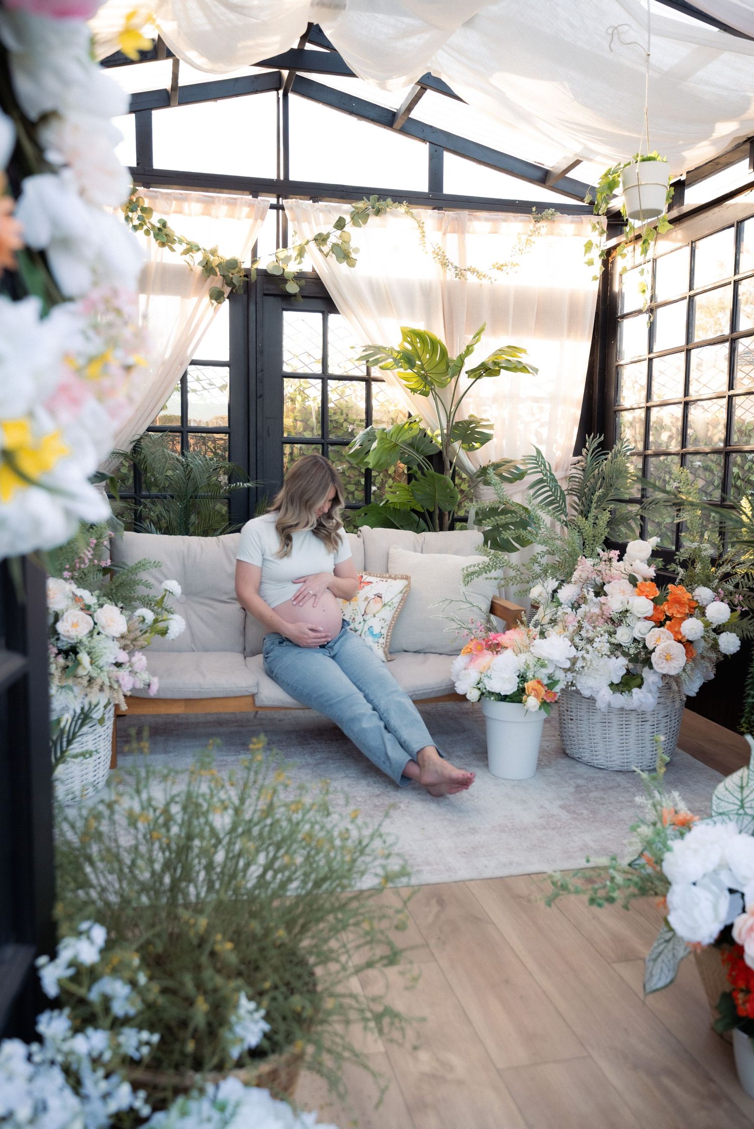 A modern greenhouse sunroom decorated with white flowers and plants features hardwood floors and black-framed windows.