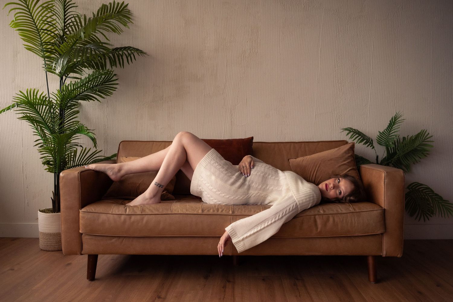 Person reclining on a brown leather sofa with green palm plants in the background.