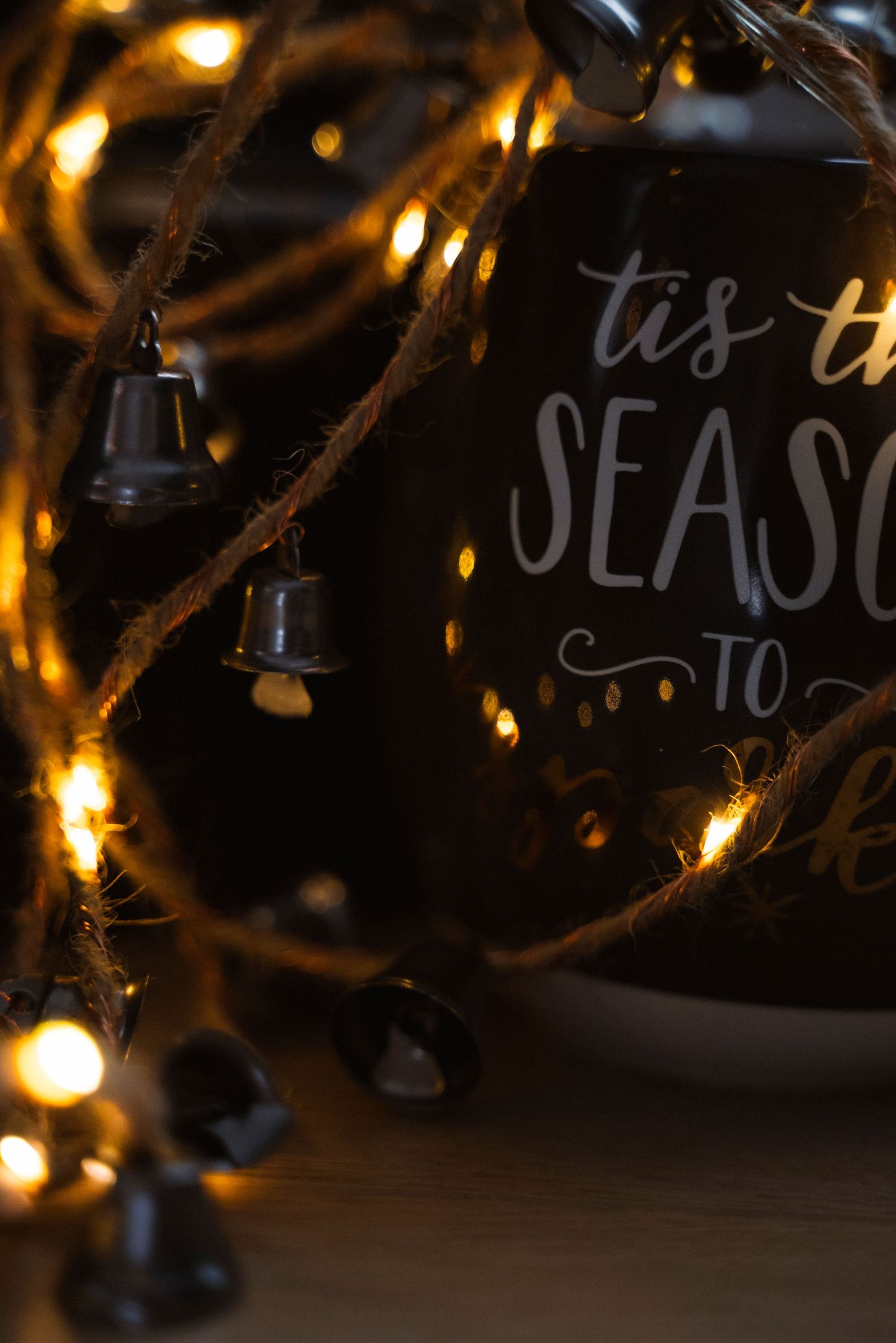 Close-up of a holiday ornament with 'tis the season' text surrounded by warm glowing Christmas lights against dark background.