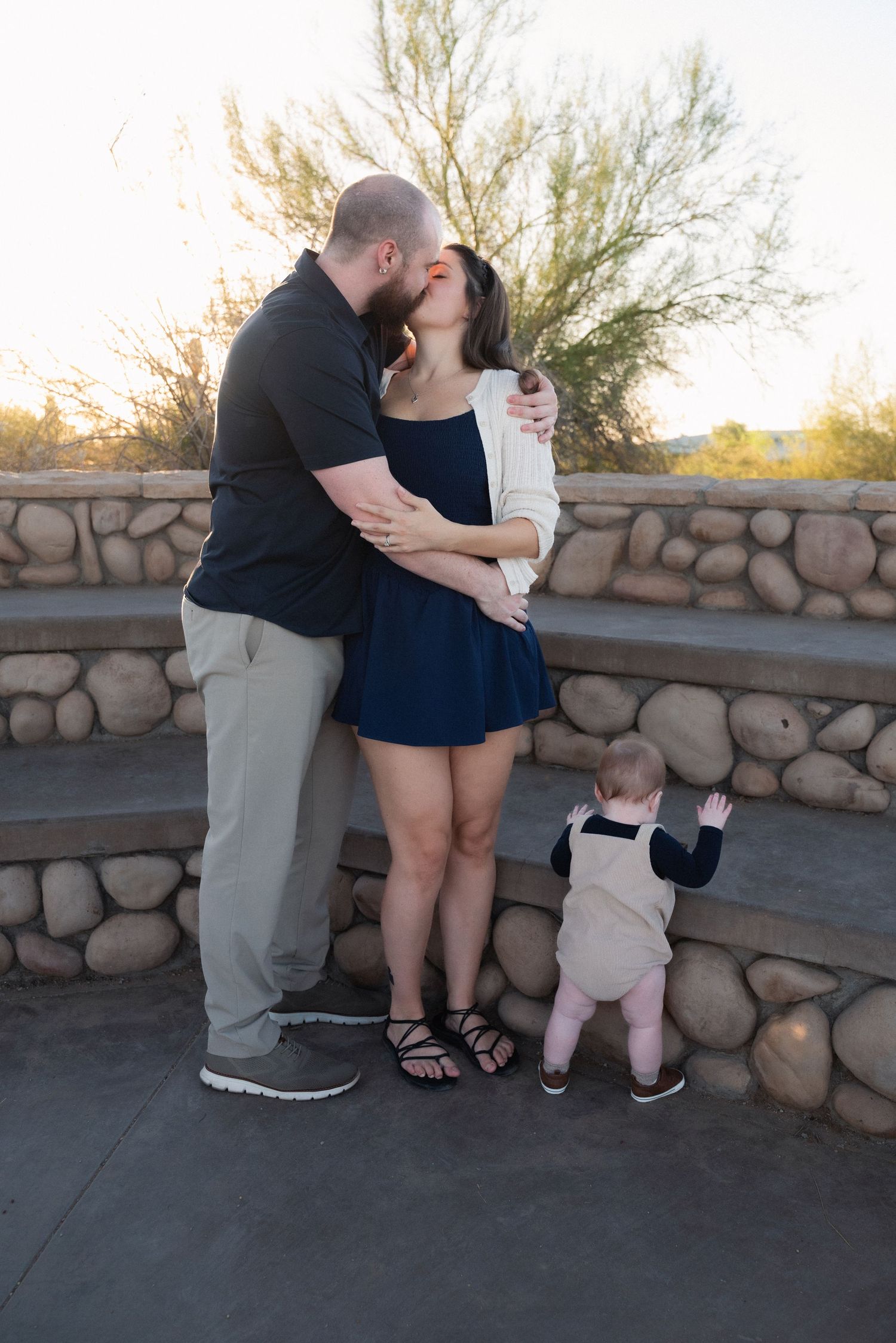 A couple shares a tender moment by a stone wall at sunset while their toddler stands nearby.