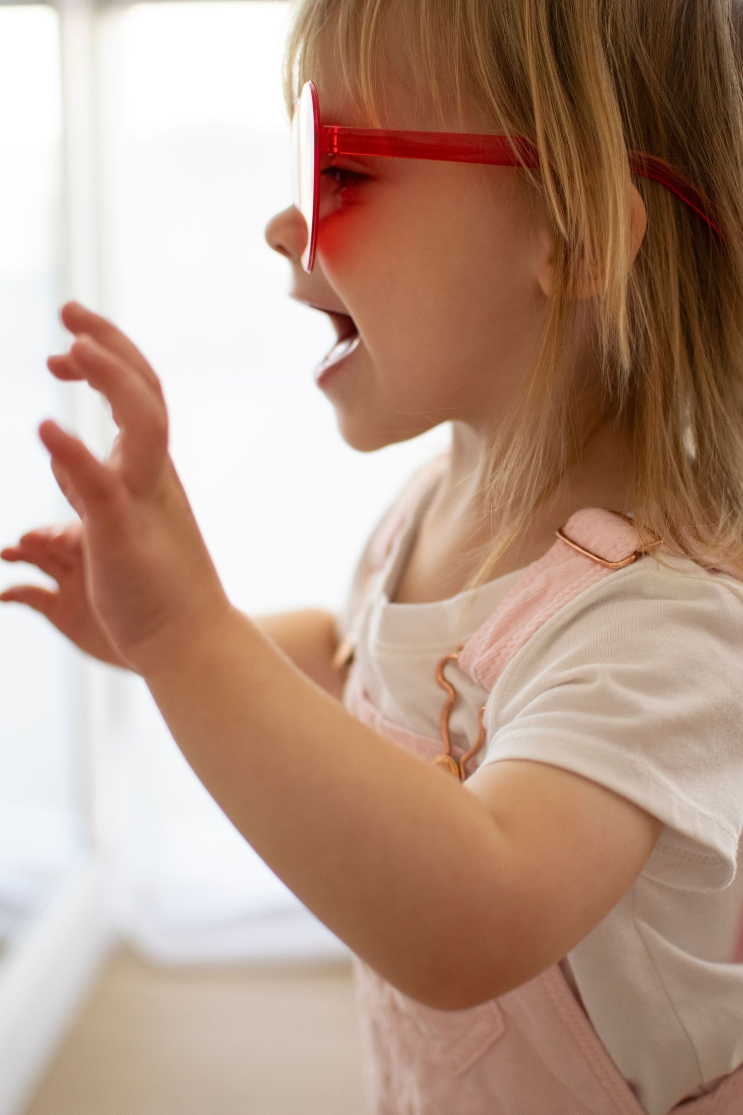 Child in red sunglasses and pink overalls gestures playfully near a window.