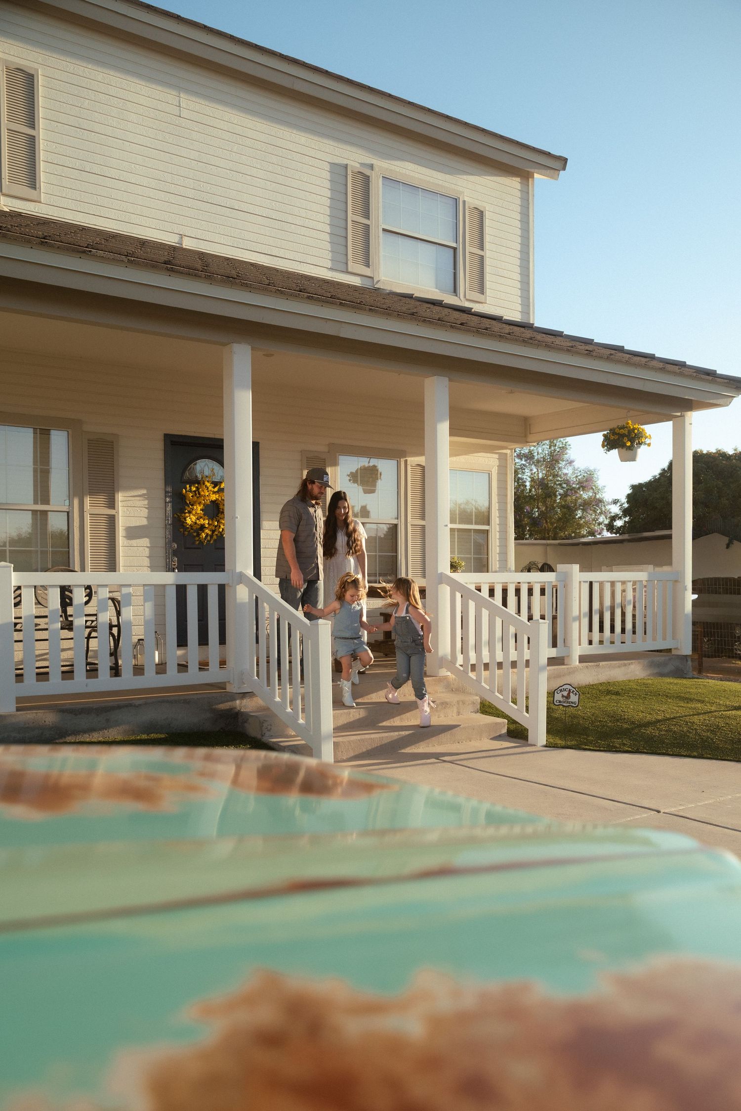 Family enjoying a sunny afternoon on the front porch of a charming two-story white farmhouse with wrap-around railing.