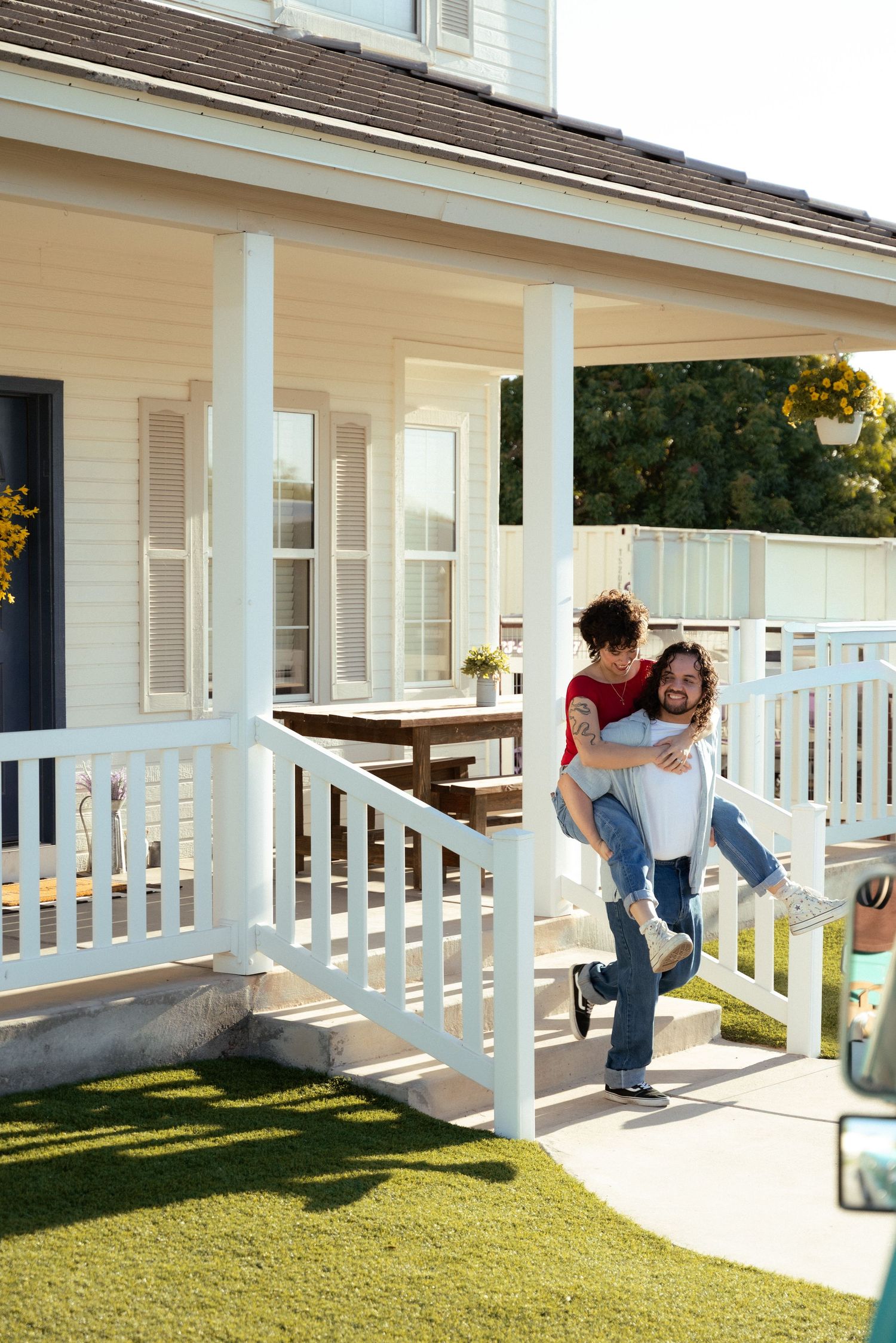 A happy couple enjoys a piggyback ride on the porch of a charming white suburban home on a sunny day.