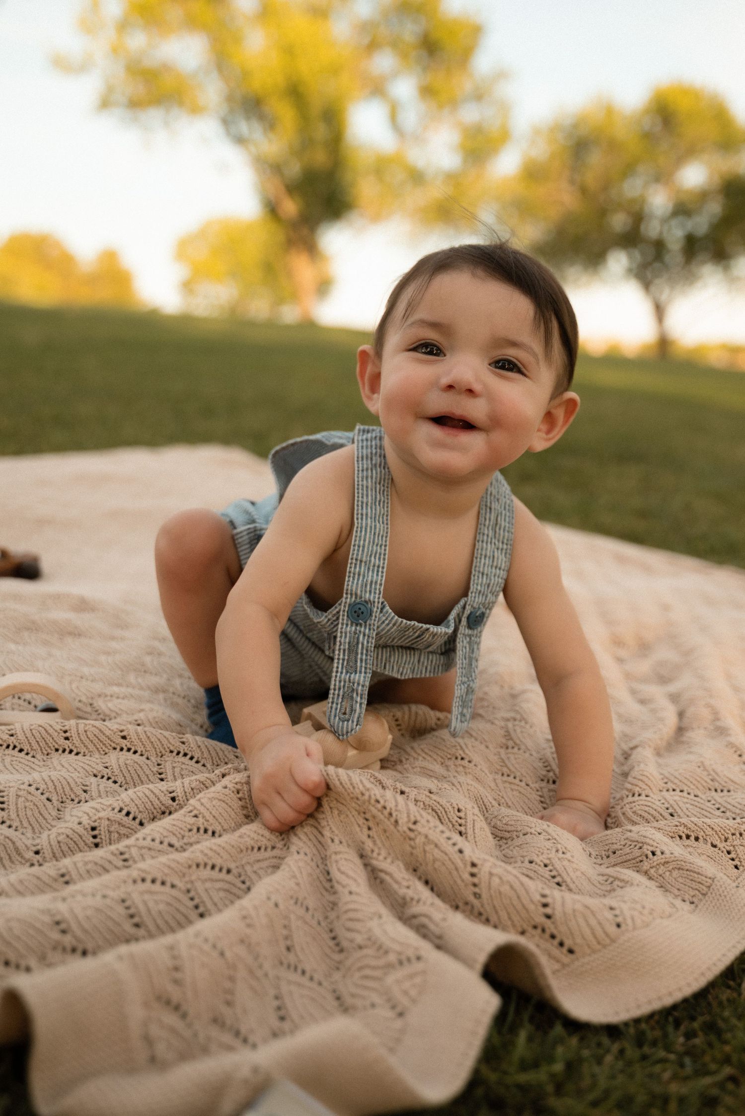 Smiling baby in gray suspender overalls crawling on a knit blanket outdoors in a sunny green park.