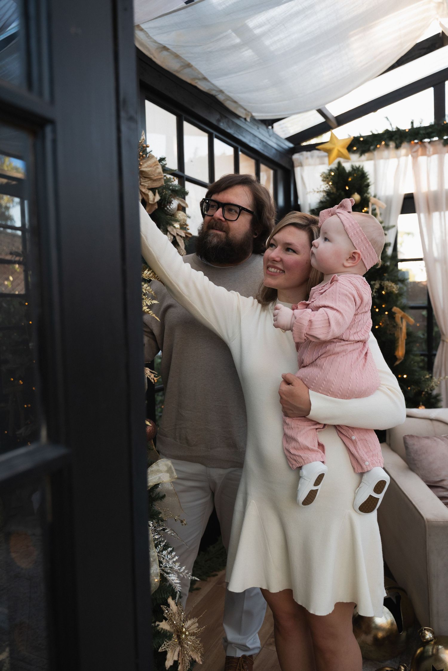 A family decorates a Christmas tree in a cozy greenhouse, with a man, woman in white dress, and baby in pink.