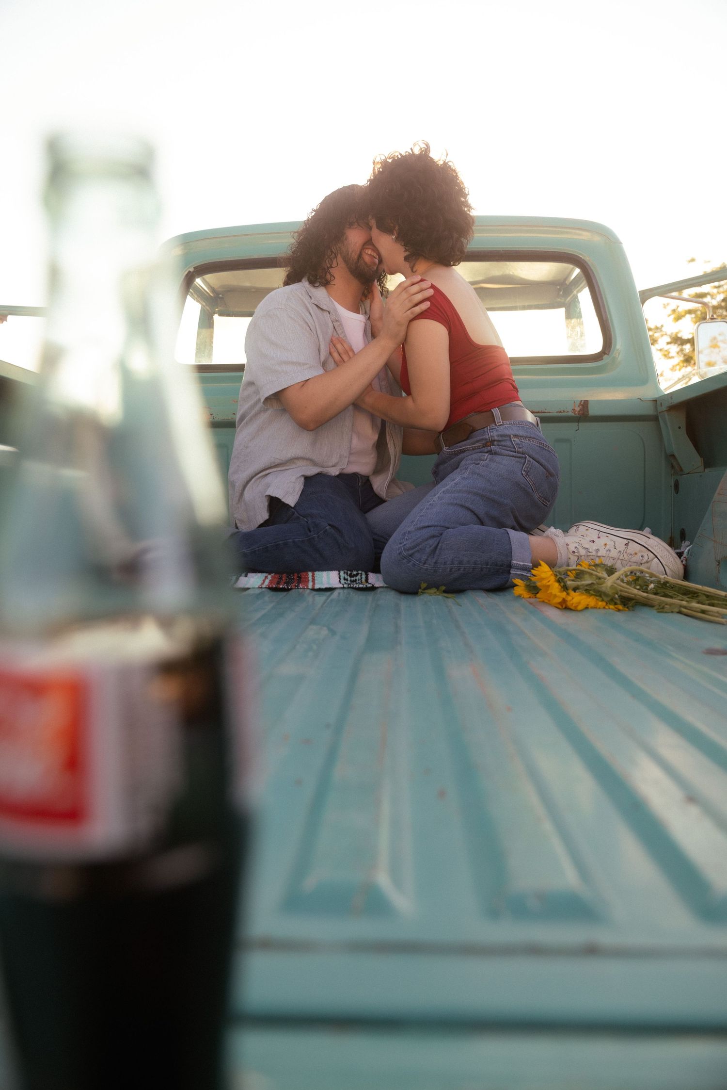 A romantic couple kissing in the bed of a vintage teal pickup truck with sunflowers and soda bottles.