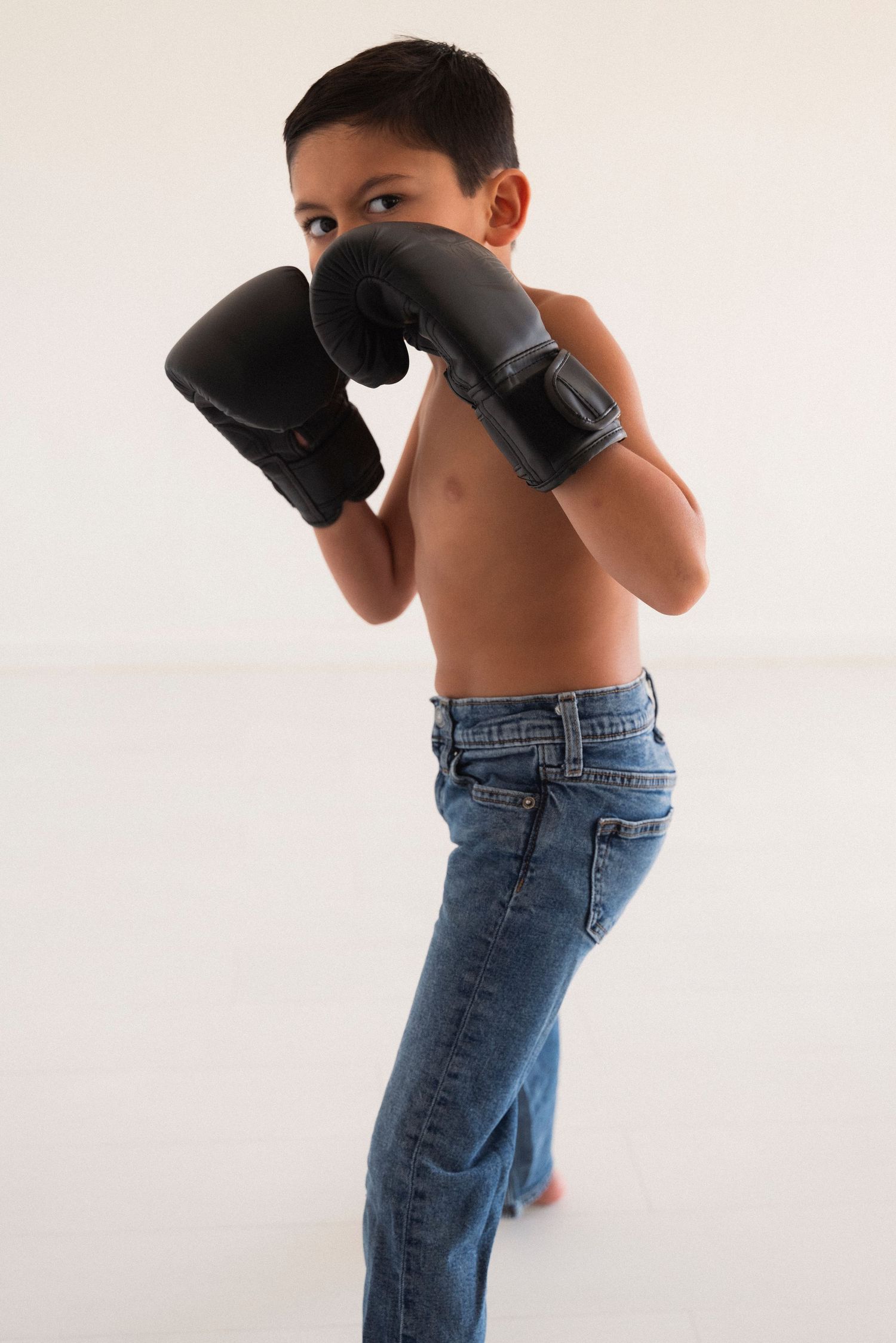 Young shirtless child in jeans strikes boxing pose with black gloves against light background.