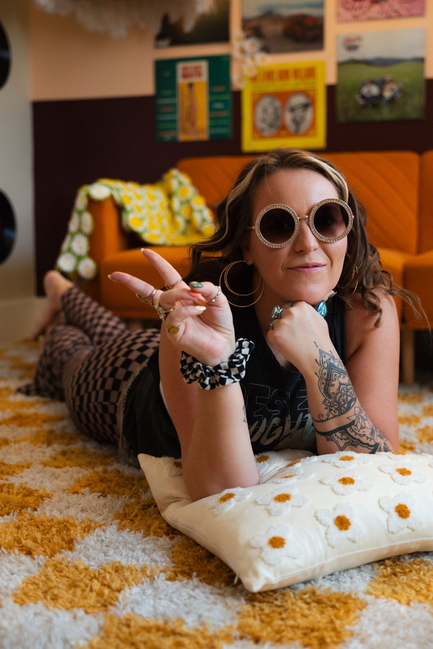 Person wearing sunglasses and bracelets relaxes on orange shag carpet with polka dot pillow in retro-styled room.