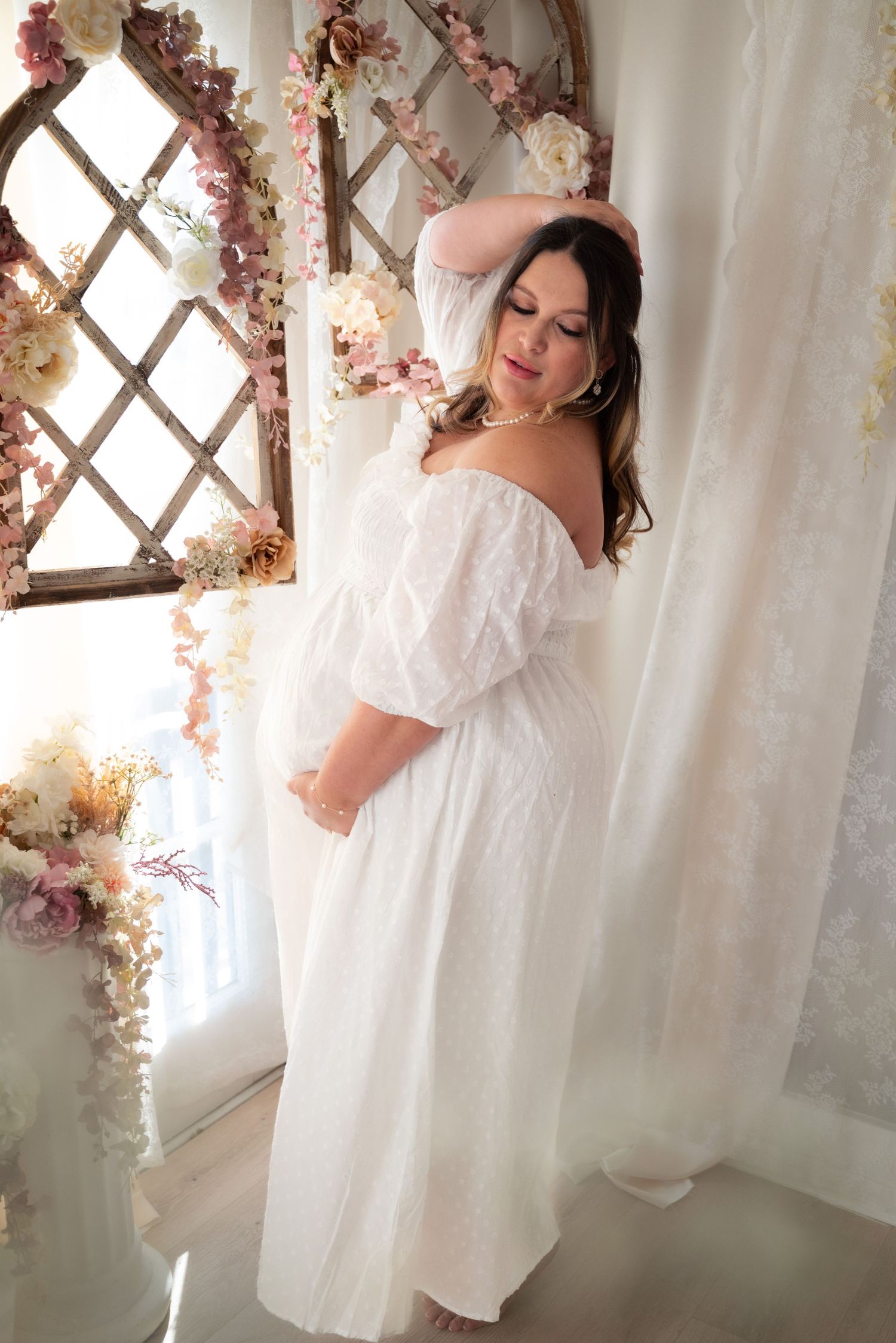 Ethereal boudoir photoshoot with soft white curtains and pink floral accents creating a dreamy romantic atmosphere.
