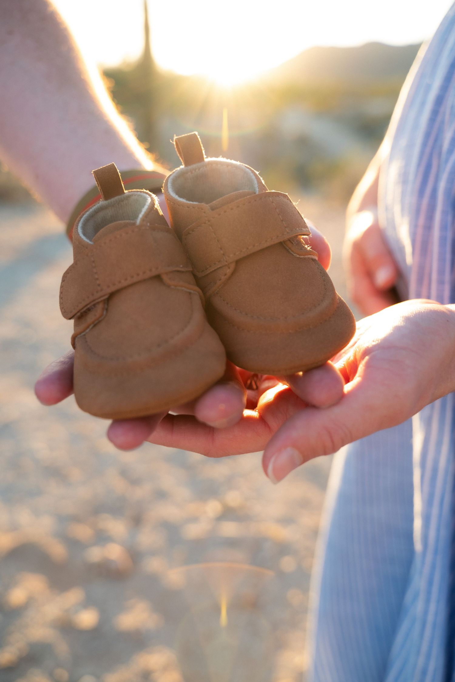 Hands holding tiny brown baby shoes against a sunset-lit beach background.