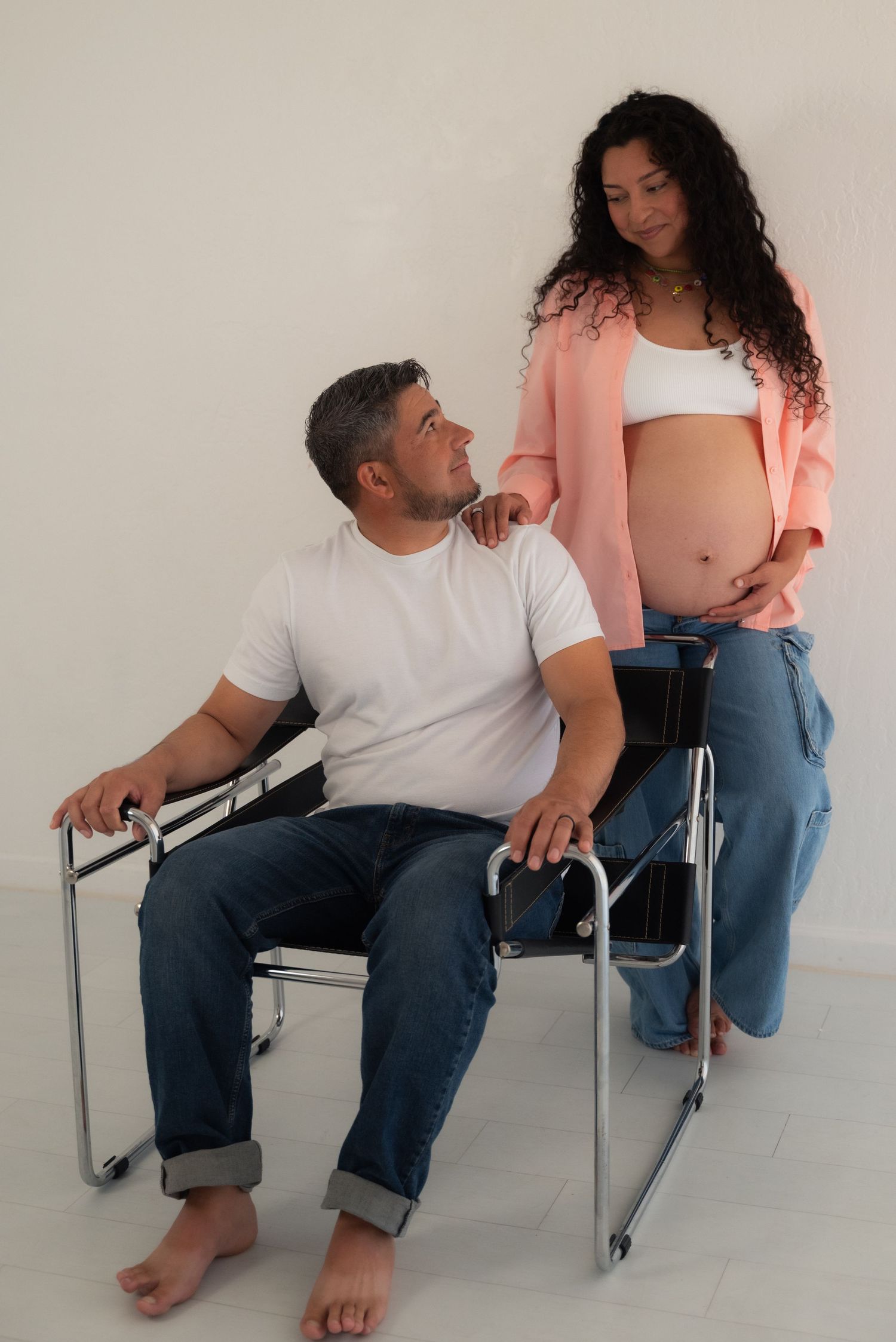 A series of intimate maternity photos showing a couple sitting in chairs against a white wall.