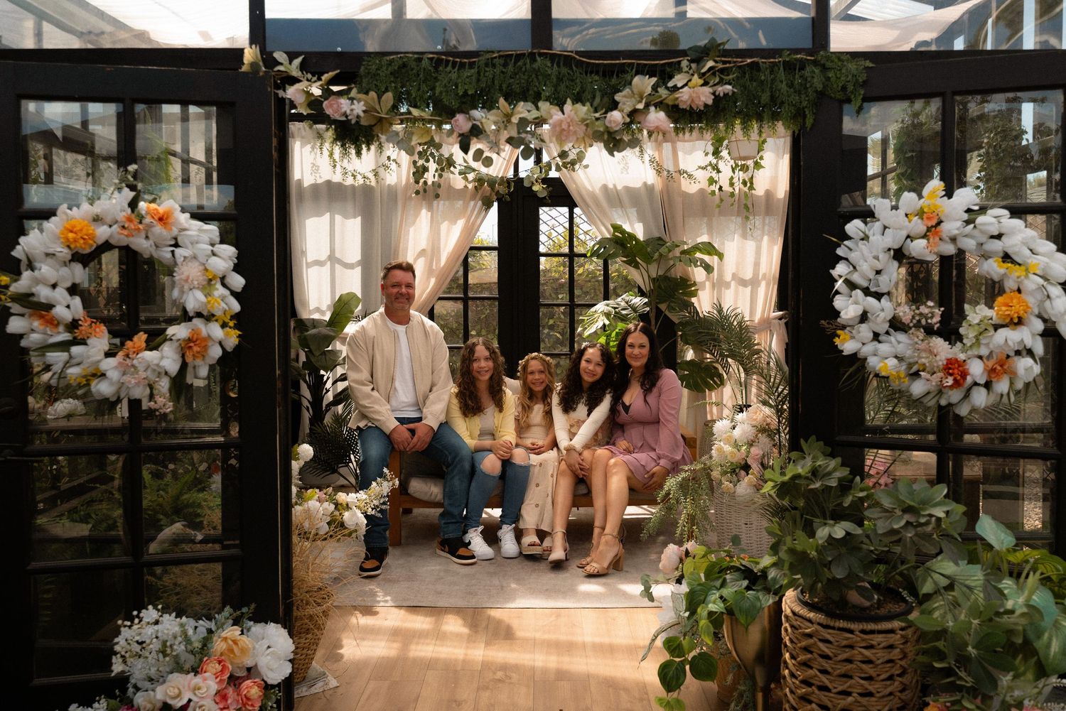 Family of five posing in a floral greenhouse doorway surrounded by colorful flower arrangements and lush greenery.