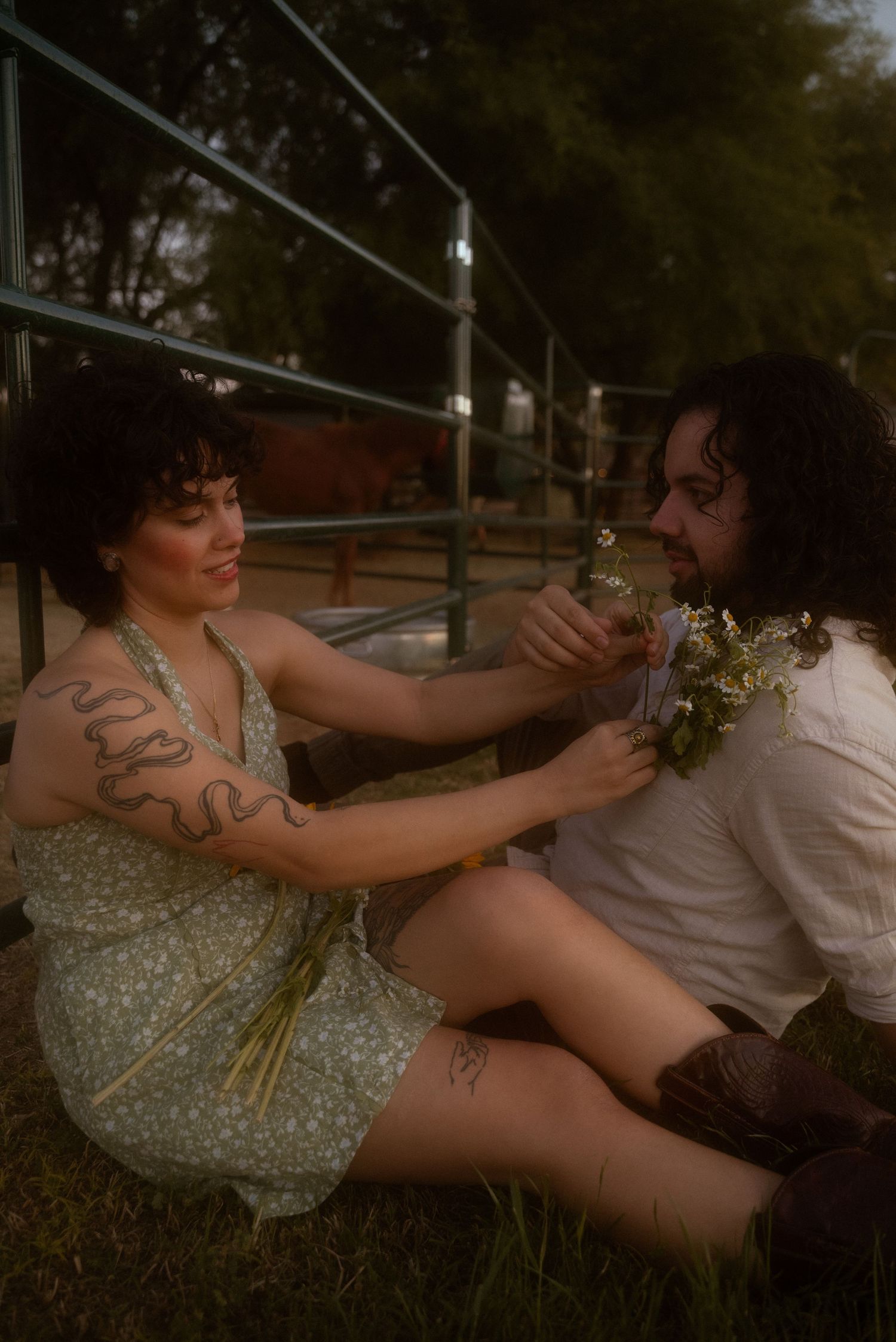 A woman with tattoos in a floral dress shares wildflowers with a curly-haired man sitting on grass outdoors.