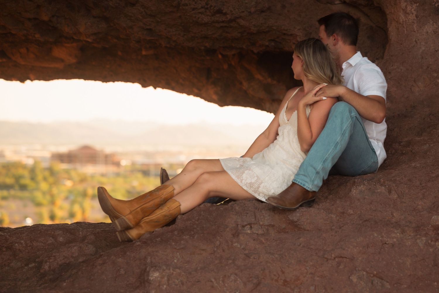 Couple sitting together in a red rock cave opening, overlooking a scenic desert landscape at sunset.