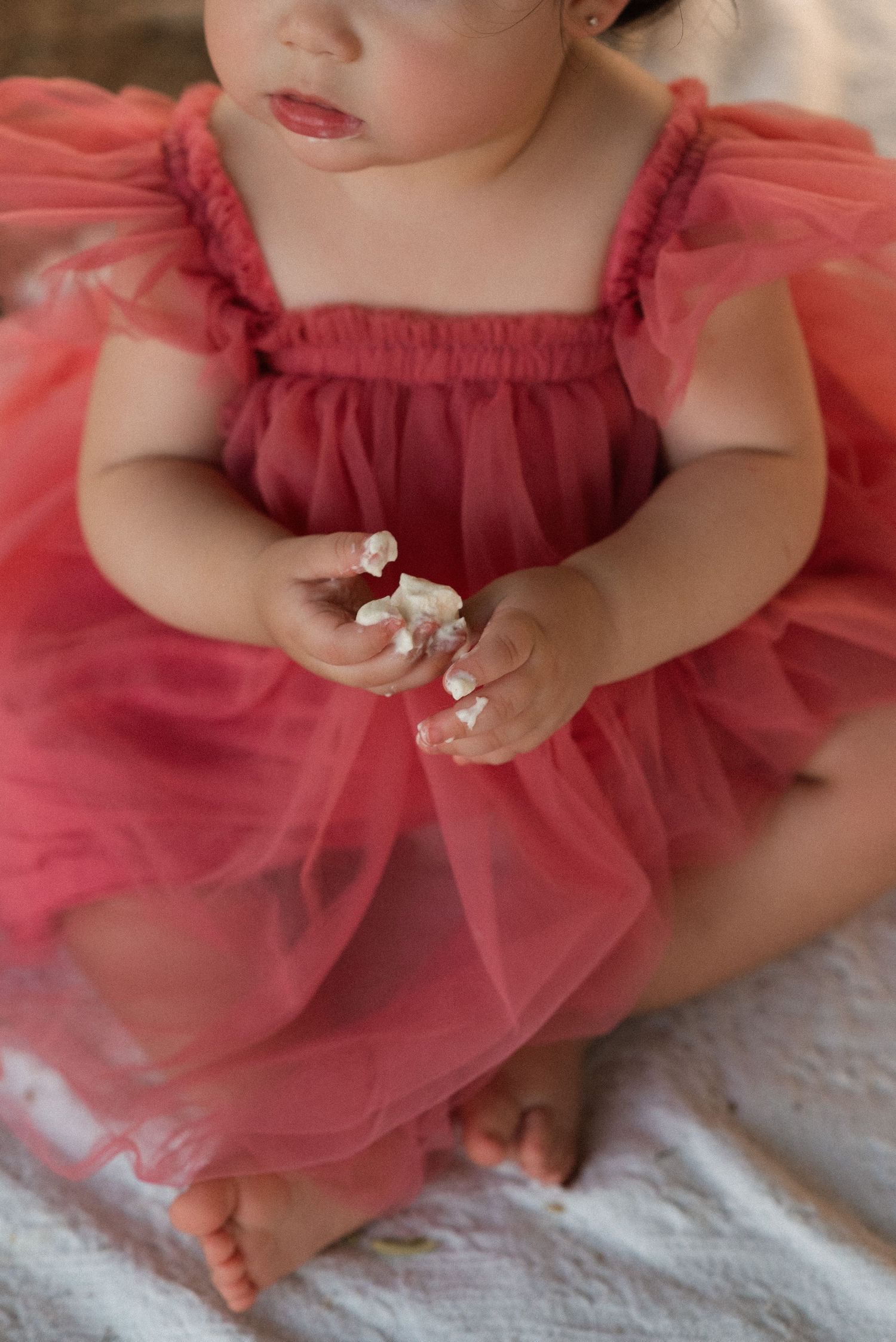 Baby girl in a pink tulle dress sits holding cake during a first birthday smash cake photoshoot.