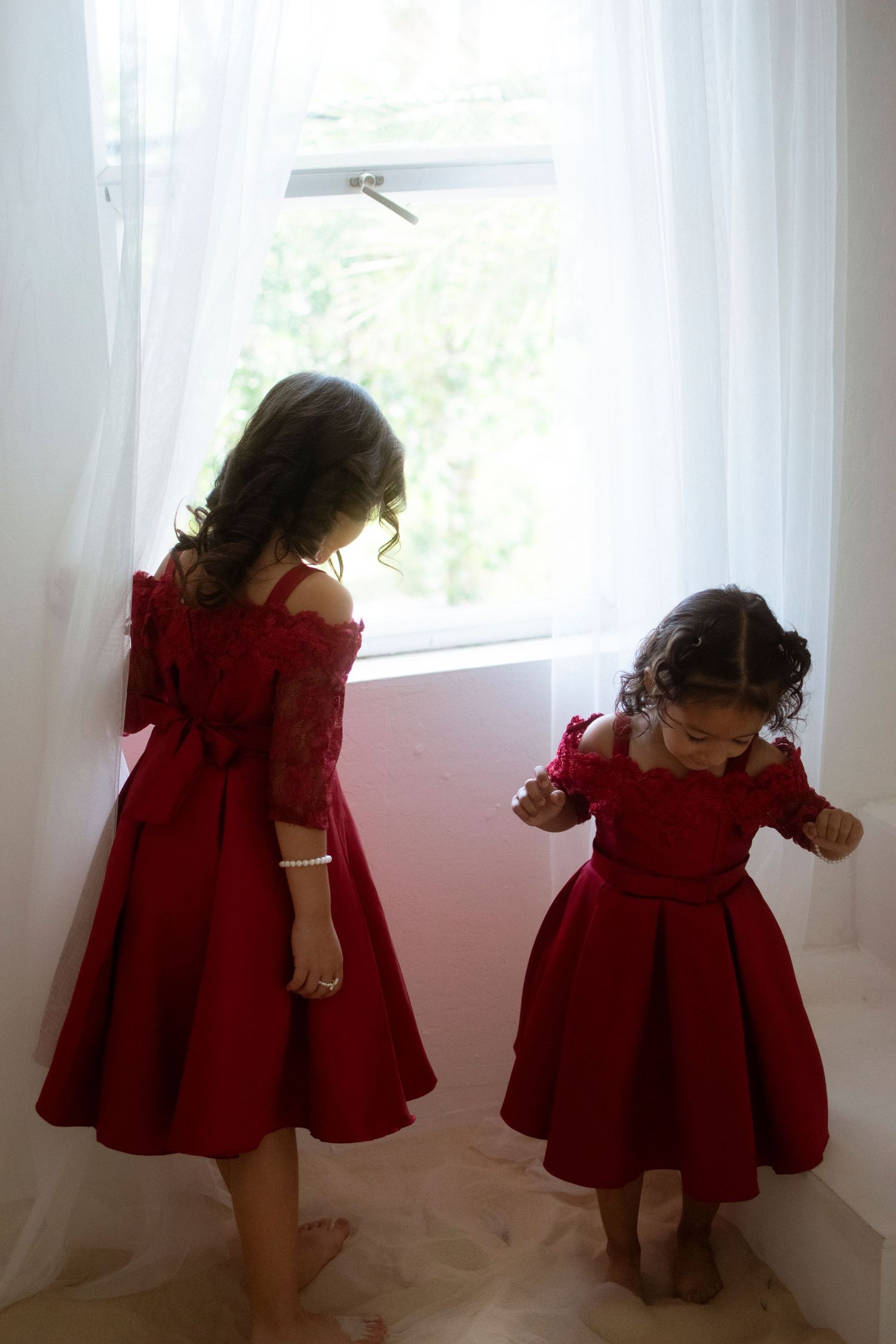 Two matching red dresses twirling by a bright window with flowing white curtains.