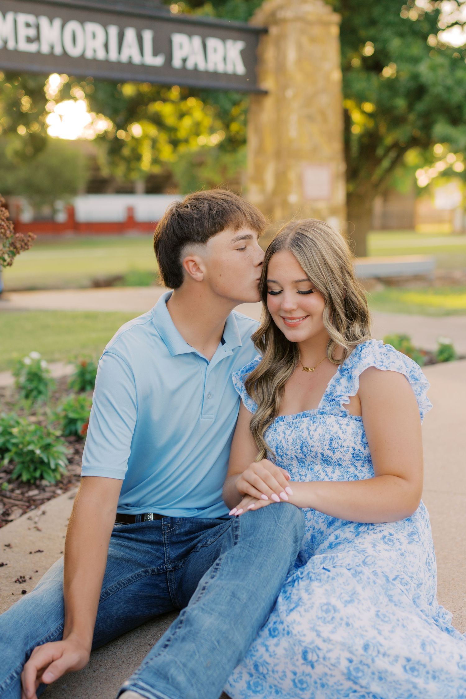 Young couple sitting at Memorial Park entrance, man kissing woman on cheek as she smiles in blue floral dress.