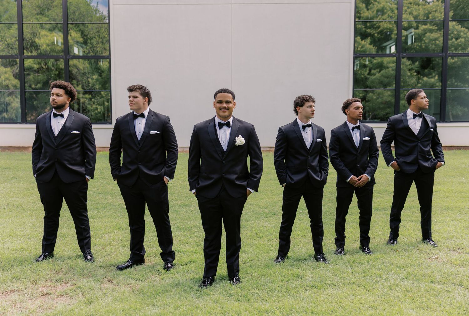 Six groomsmen in matching black tuxedos with bow ties pose outdoors on green grass at a wedding. Oklahoma wedding photographer, photographed