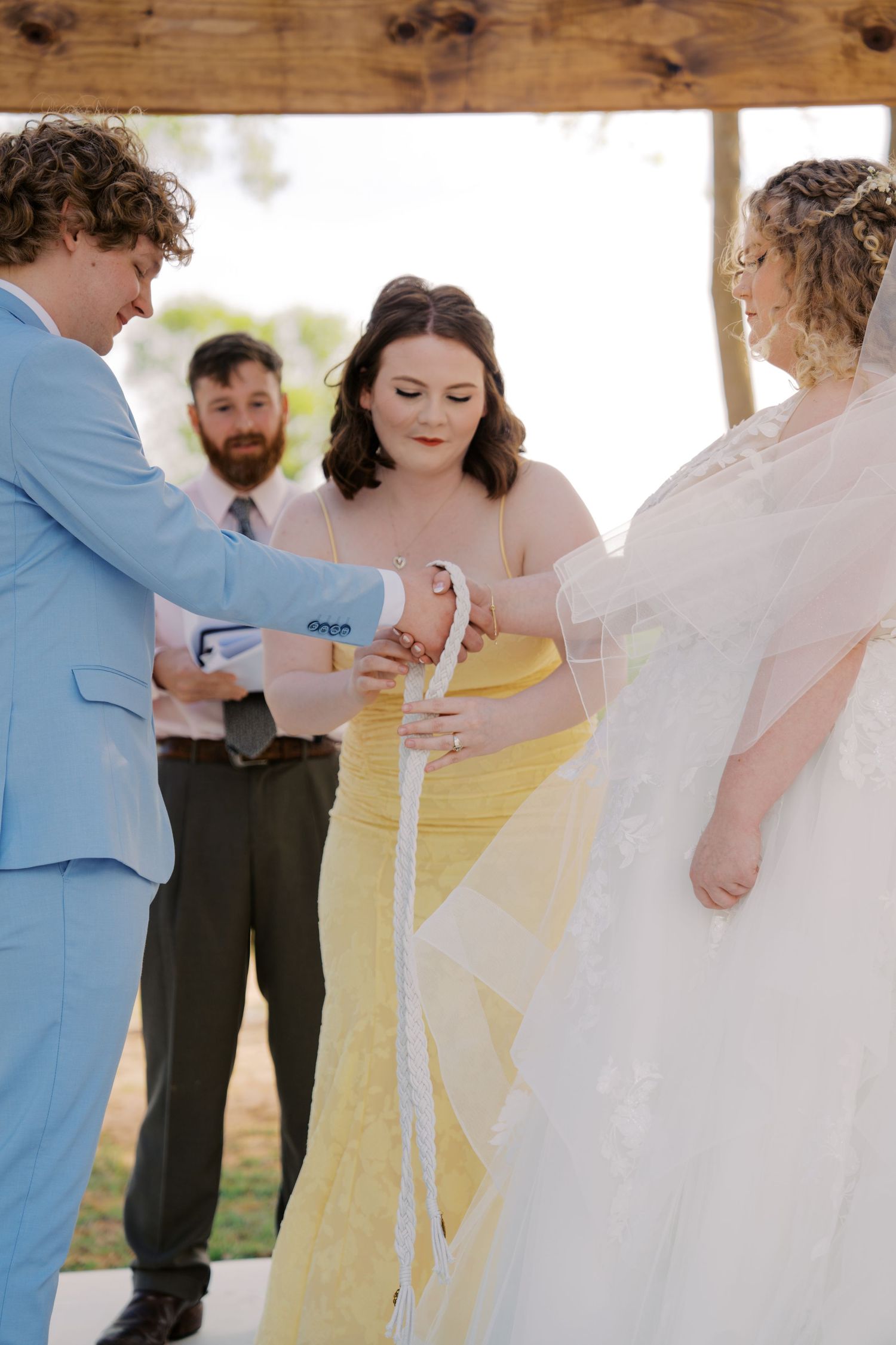 Wedding ceremony handfasting ritual with bride in white gown, groom in blue suit, and bridesmaid in yellow dress.
