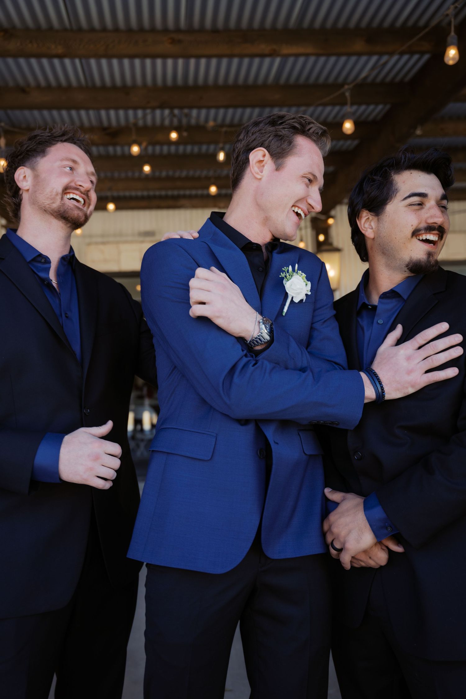 Three men in blue and black suits laughing together at a wedding, with the groom wearing a boutonniere. Oklahoma wedding photographer.
