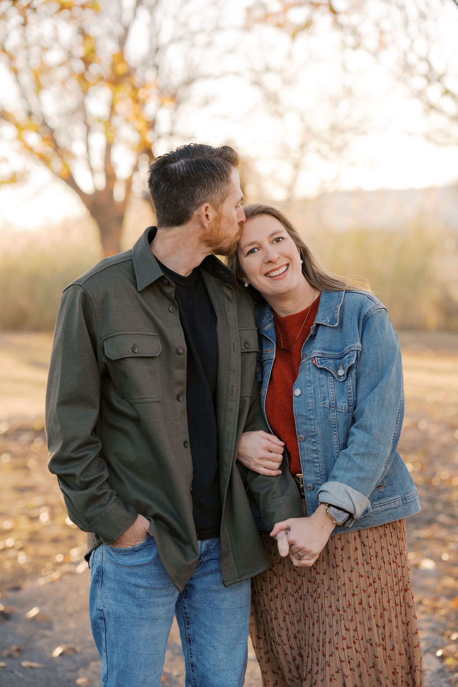 A couple shares a tender moment outdoors in autumn, man kissing woman's cheek as she smiles warmly.