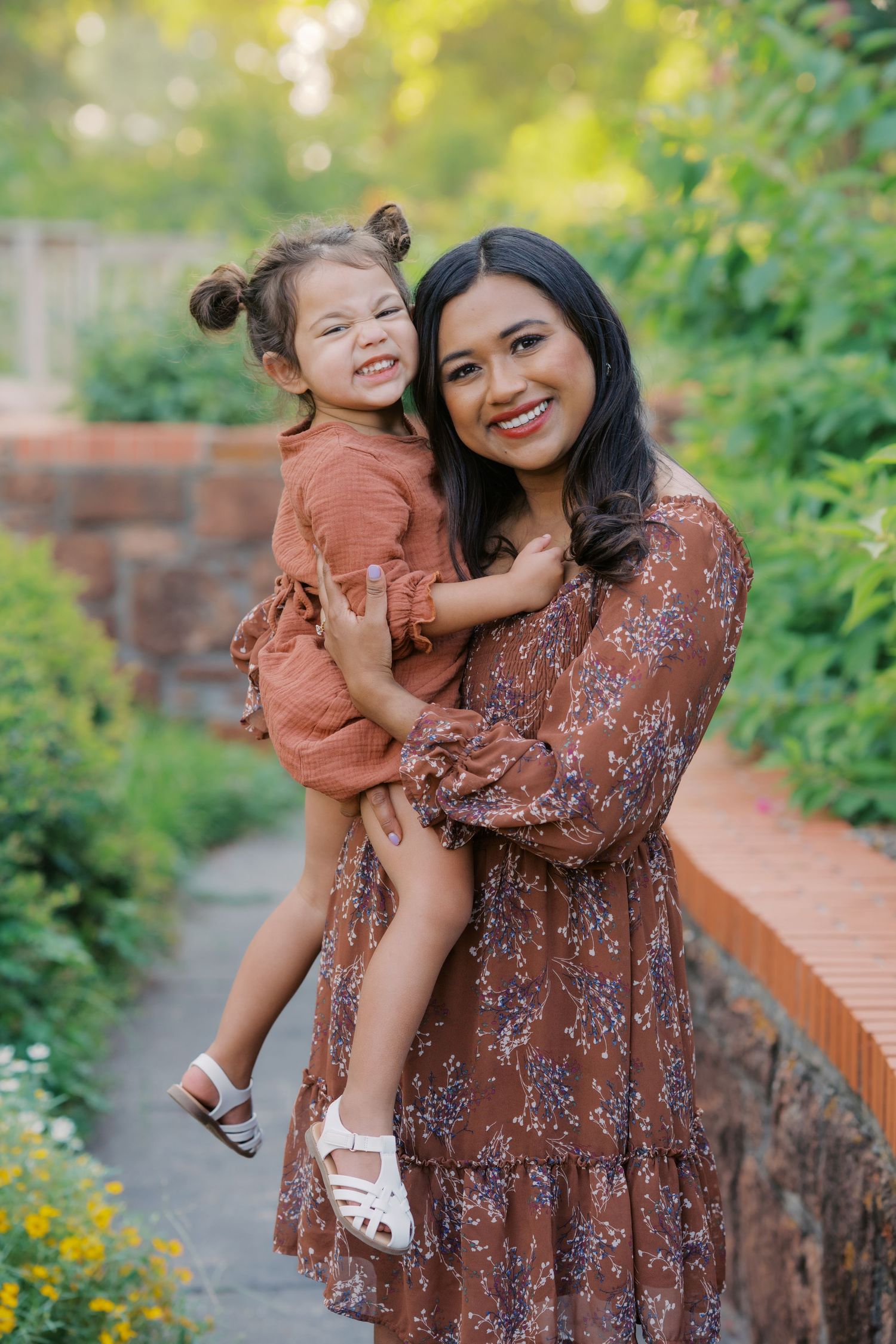 A smiling woman in a brown floral dress holds a happy toddler girl in a rust outfit outdoors in a lush garden.