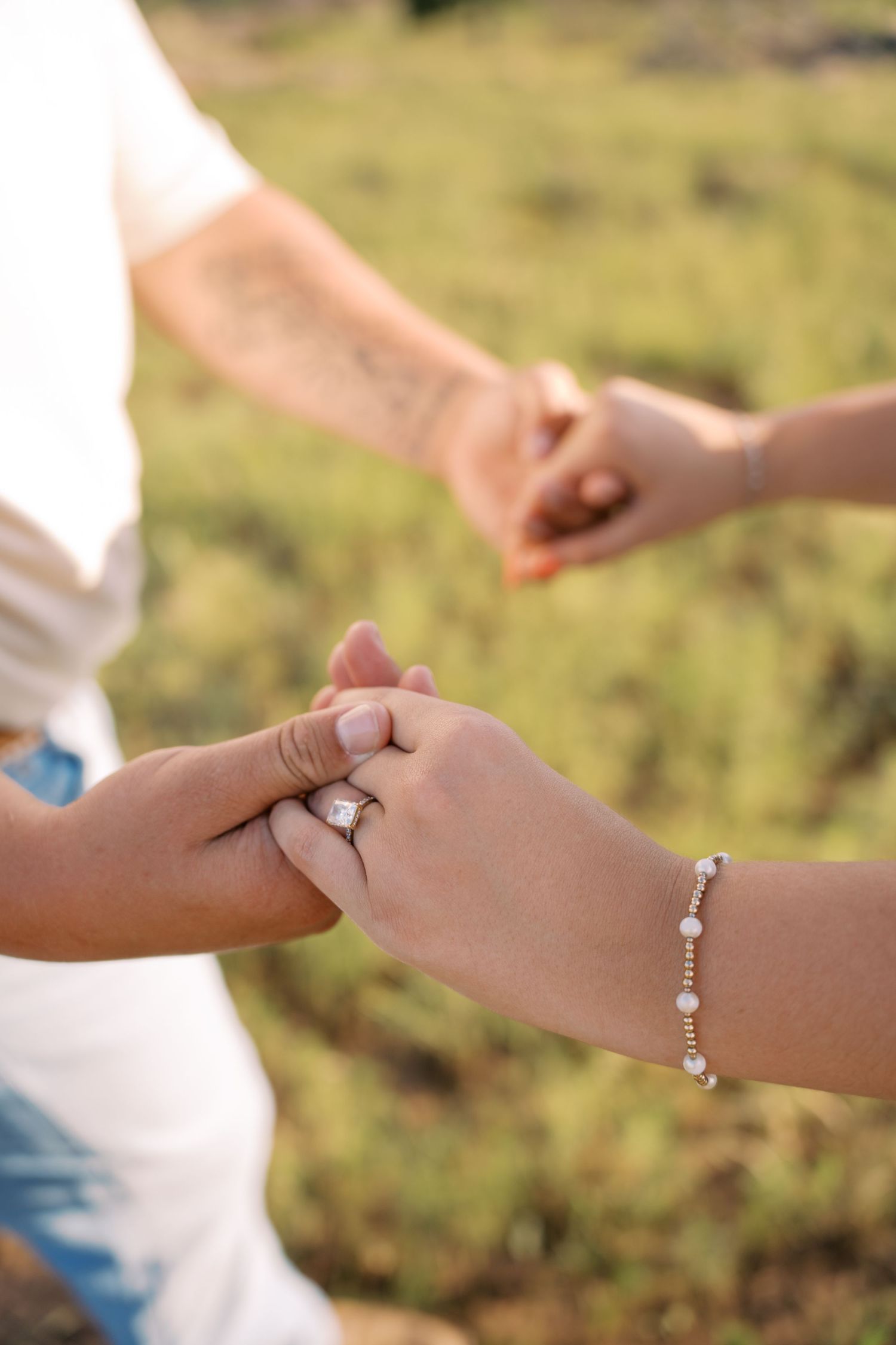 Close-up of engaged couple holding hands showing diamond engagement ring and delicate pearl bracelet.