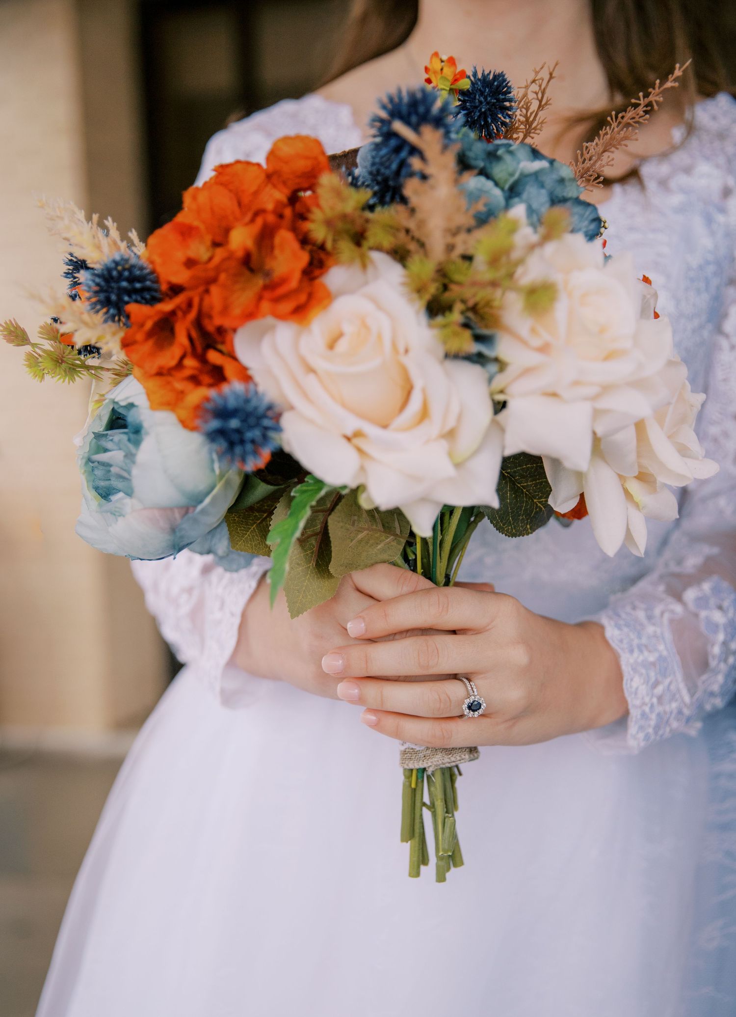 Bride holding colorful bouquet with orange, blue, and cream flowers in white lace gown.