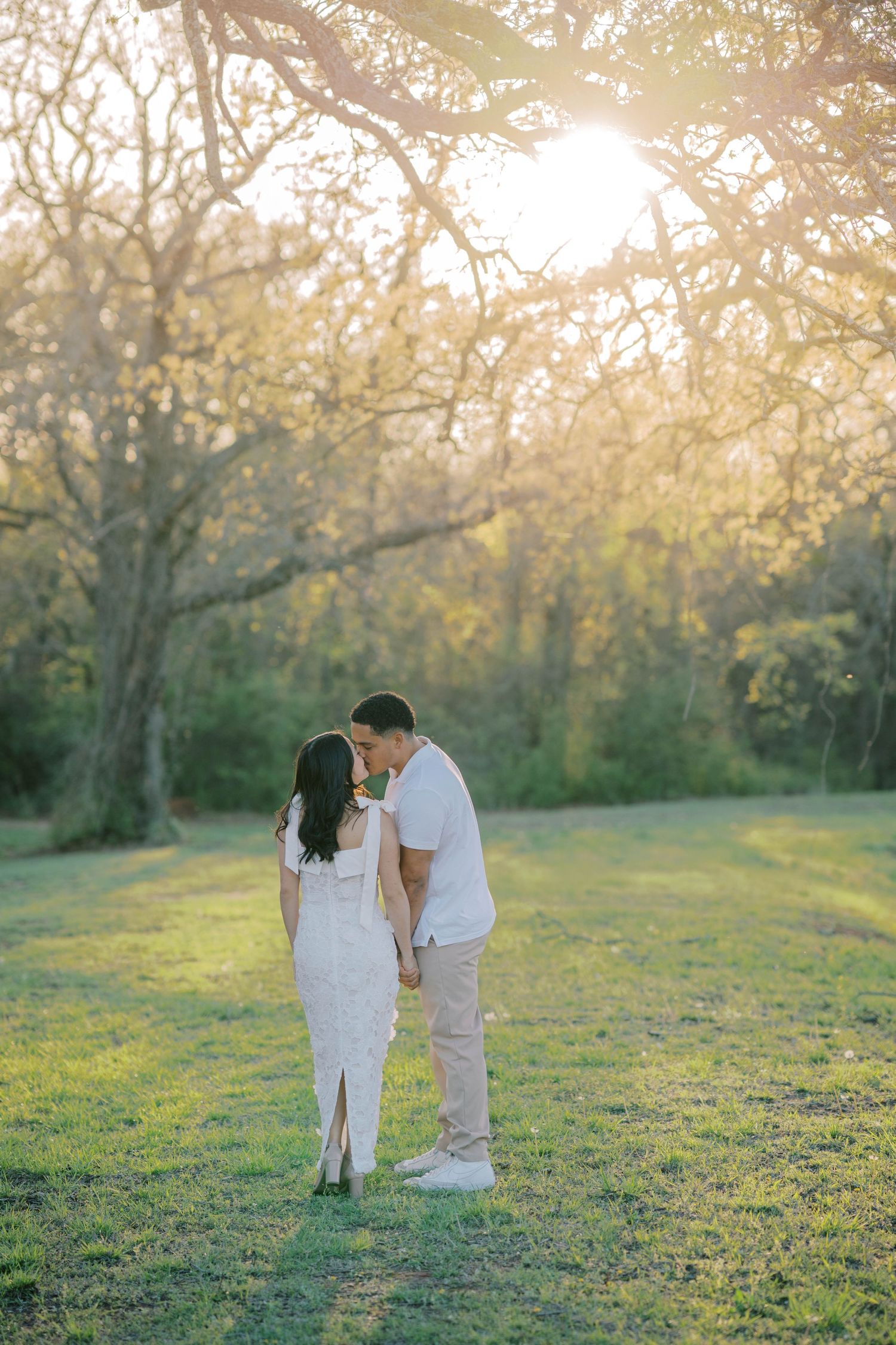 Couple shares a tender kiss while holding hands in a sunlit park surrounded by lush trees.