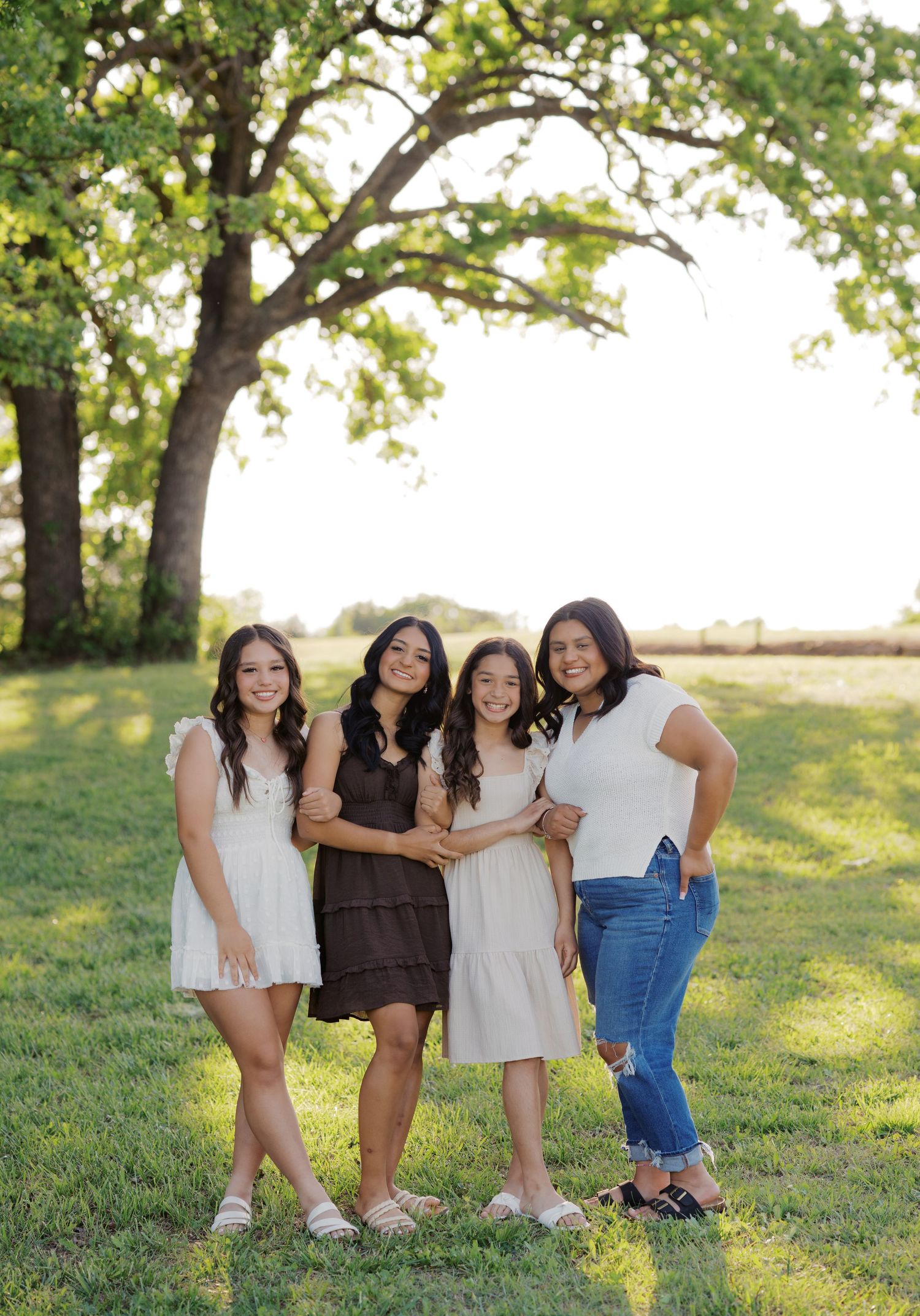Four young women smiling and posing together outdoors under a large oak tree on a sunny day.