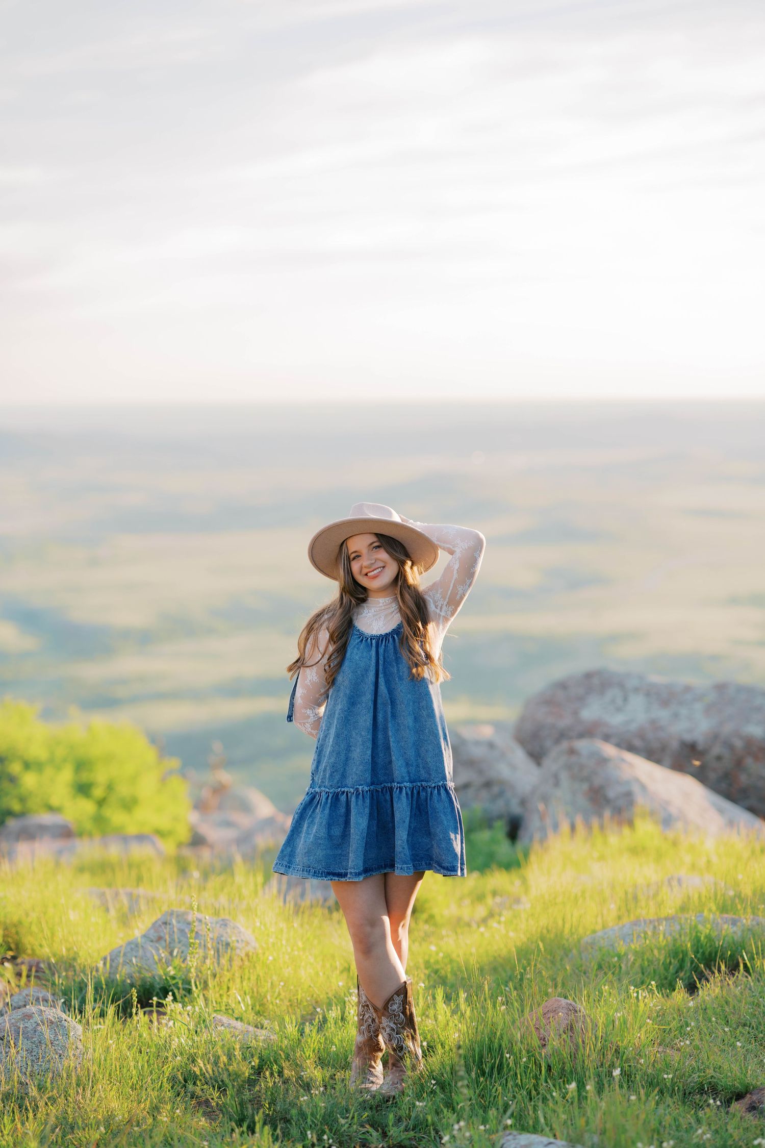 Young woman in denim dress, wide-brim hat, and cowboy boots standing on a grassy hilltop with scenic views.