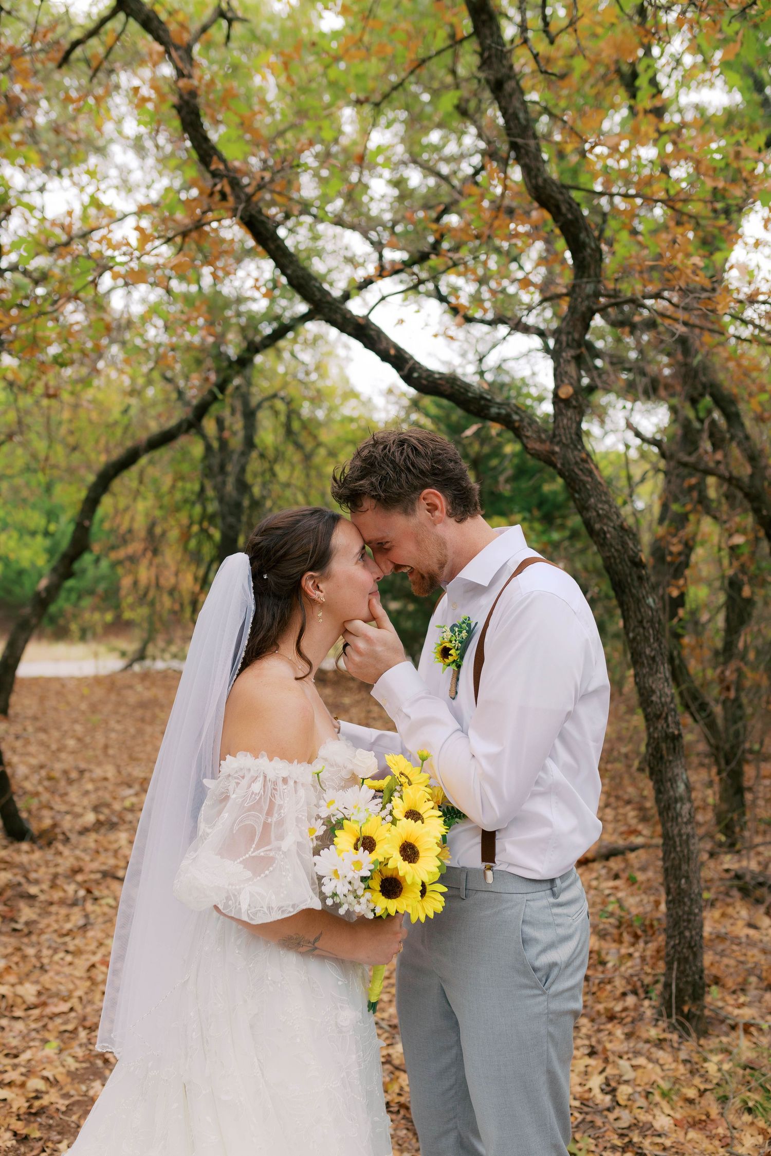 Bride and groom touch foreheads tenderly in autumn forest, groom holding sunflower bouquet. Oklahoma wedding photographer.