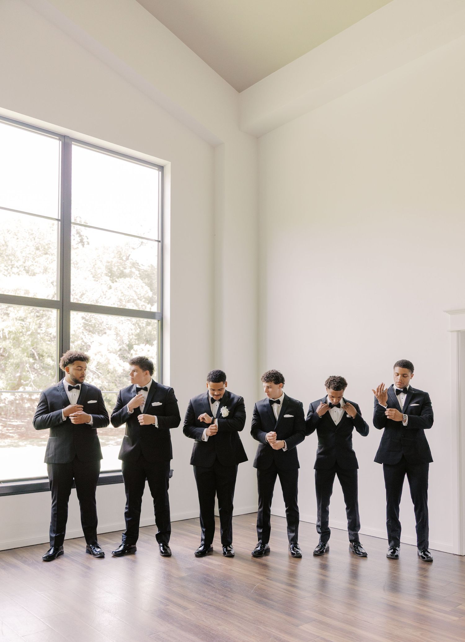 Six groomsmen in black tuxedos adjusting their bow ties before a wedding at Sorelle at Meadow Creek in Oklahoma.