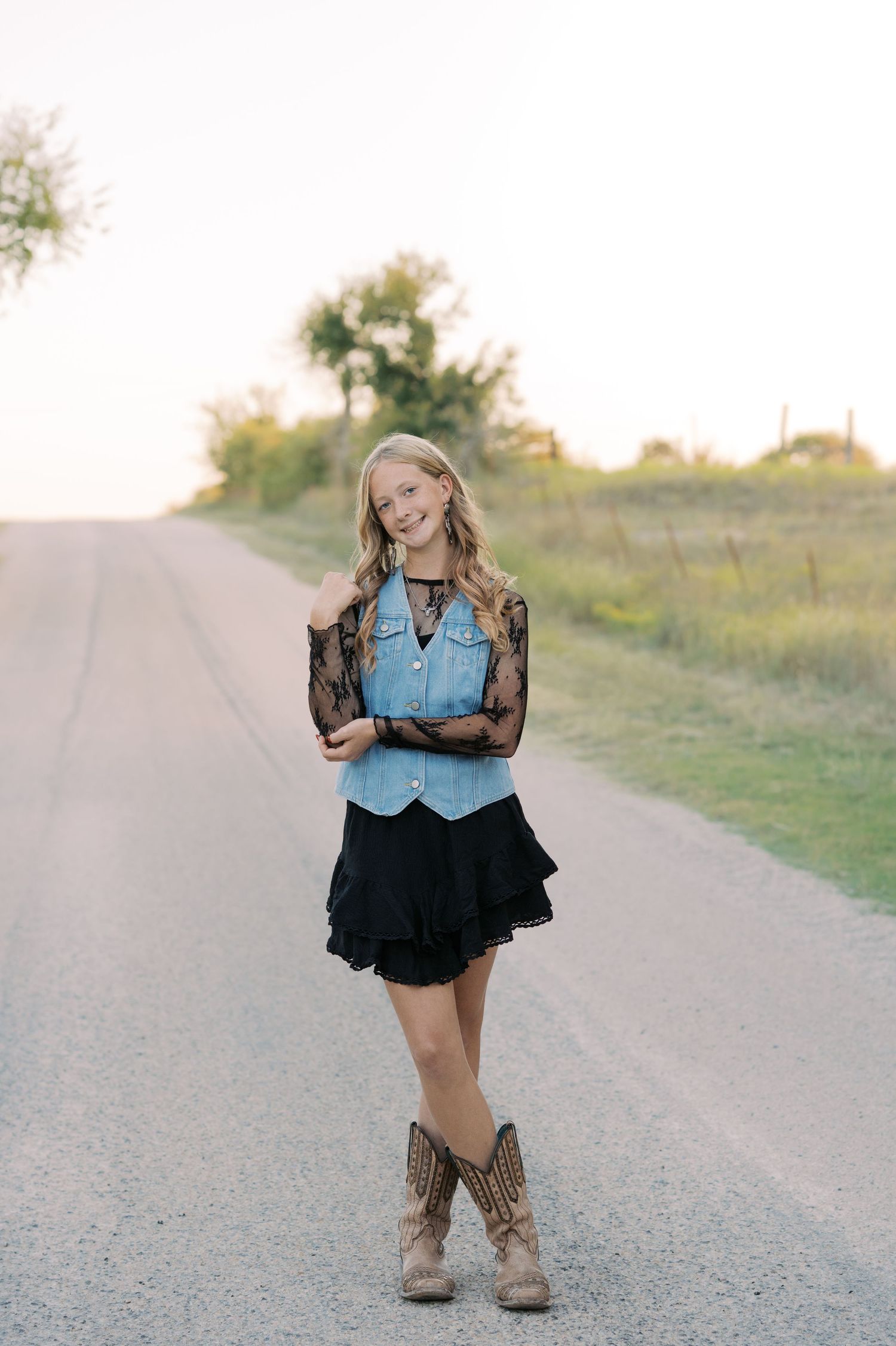 Young blonde woman in denim vest, black lace top, mini skirt, and cowboy boots posing on a rural road at sunset.