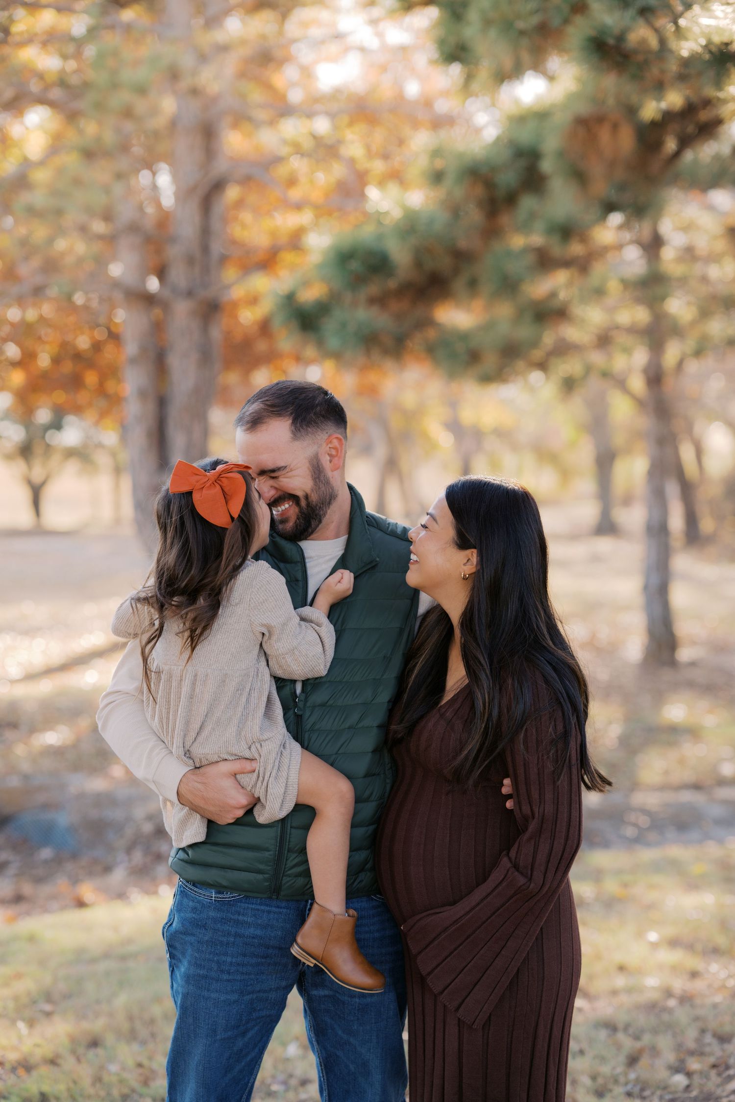 A happy family of three poses outdoors in fall, dad holding toddler with orange bow, pregnant mom smiling beside them.
