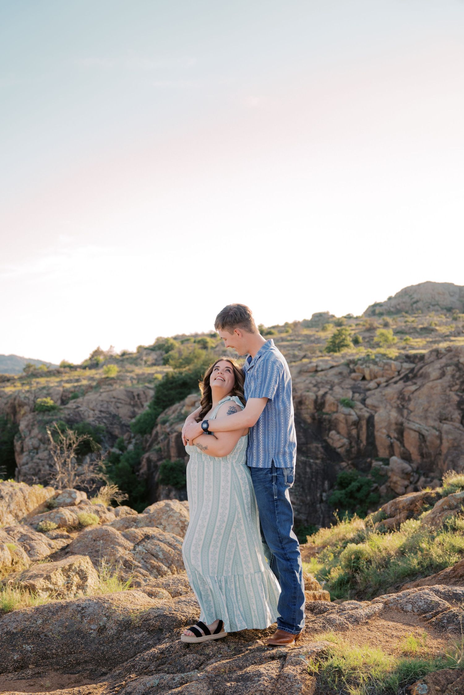 Couple embracing on rocky overlook at golden hour, woman in flowy dress smiling up at man in blue shirt and jeans.