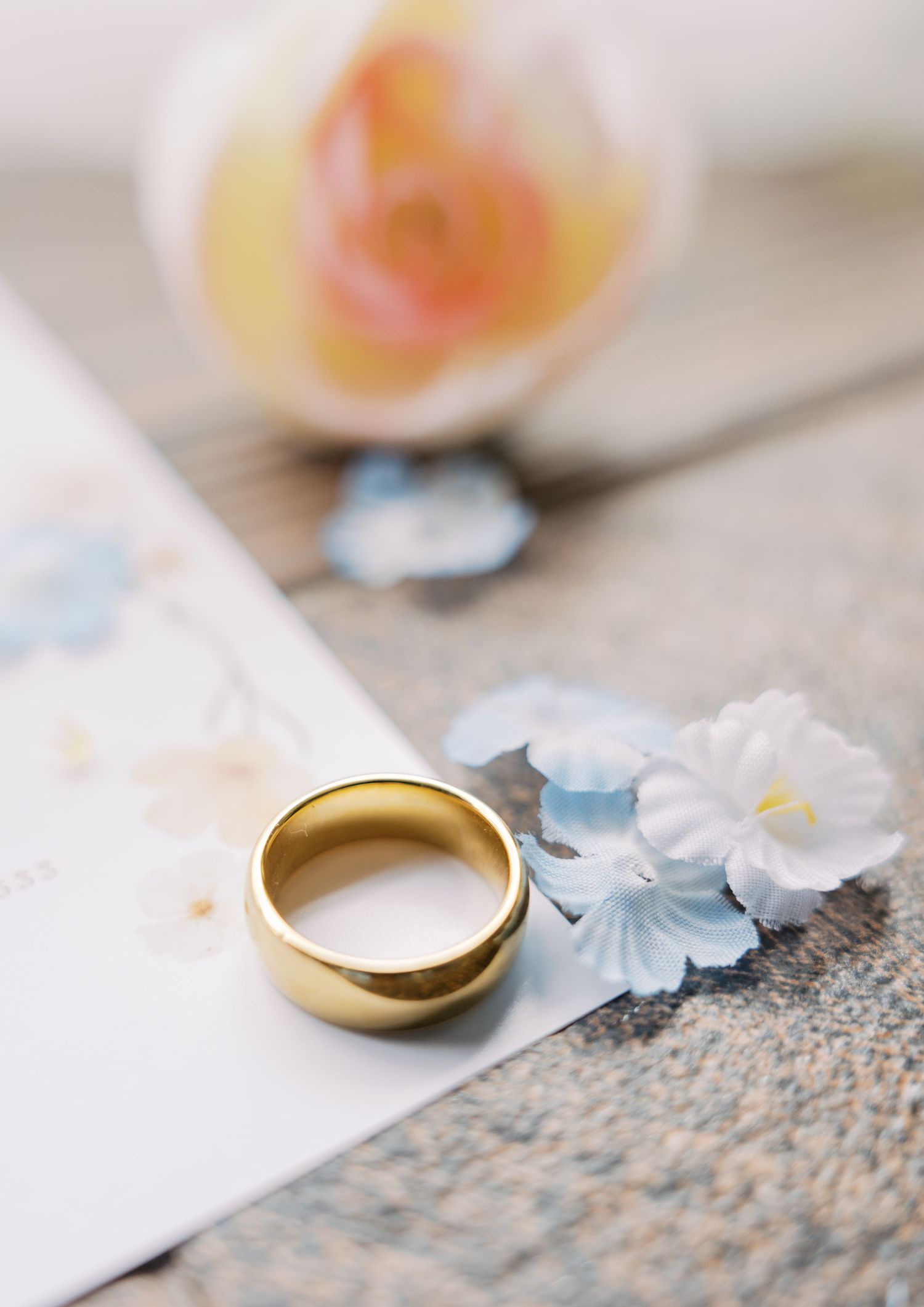Gold wedding band resting on stationery beside delicate blue flowers and a soft pink rose on a rustic wooden surface.