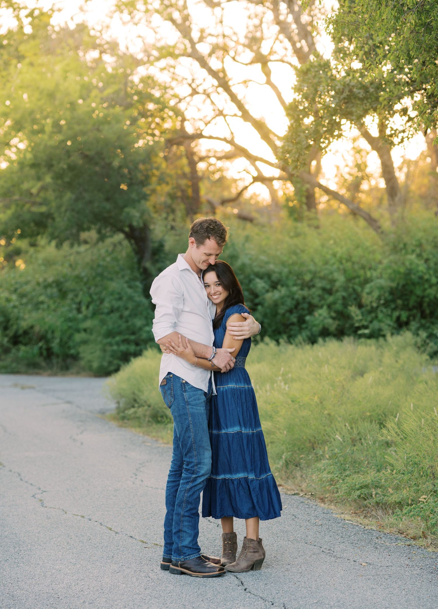 Couple embracing on a sunlit path surrounded by lush green trees during golden hour outdoors.