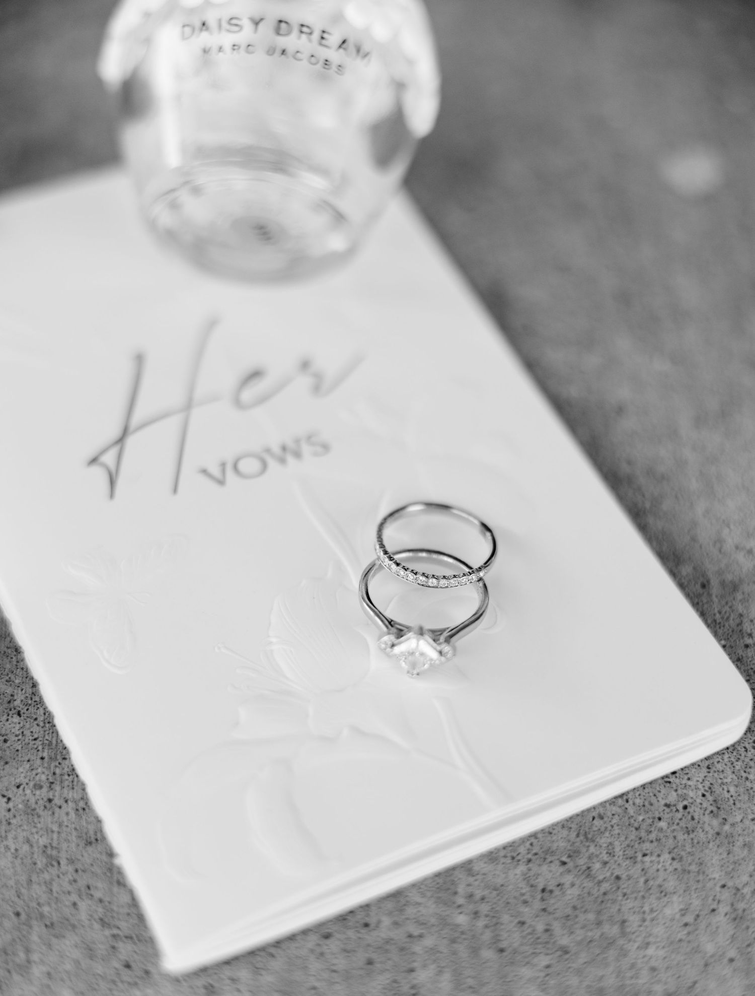 Black and white photo of wedding rings on a Her Vows book with Marc Jacobs Daisy Dream perfume bottle. Oklahoma wedding photographer.
