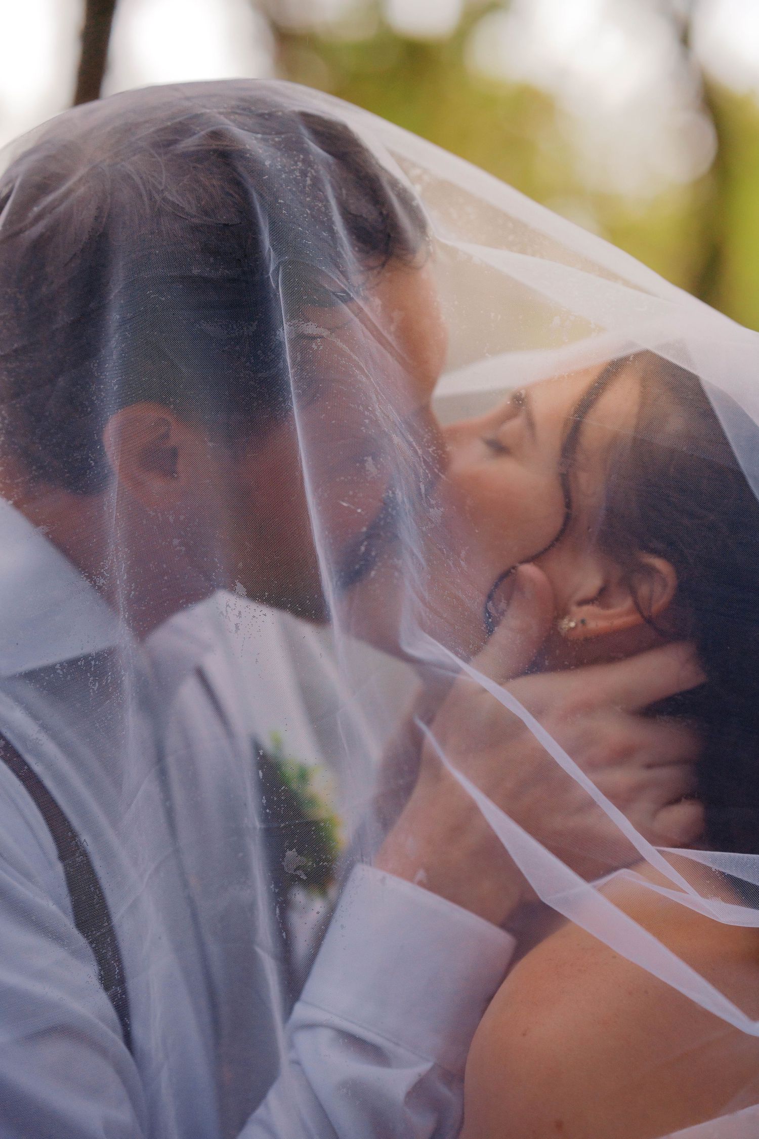 A bride and groom share a romantic kiss beneath a flowing white veil outdoors.