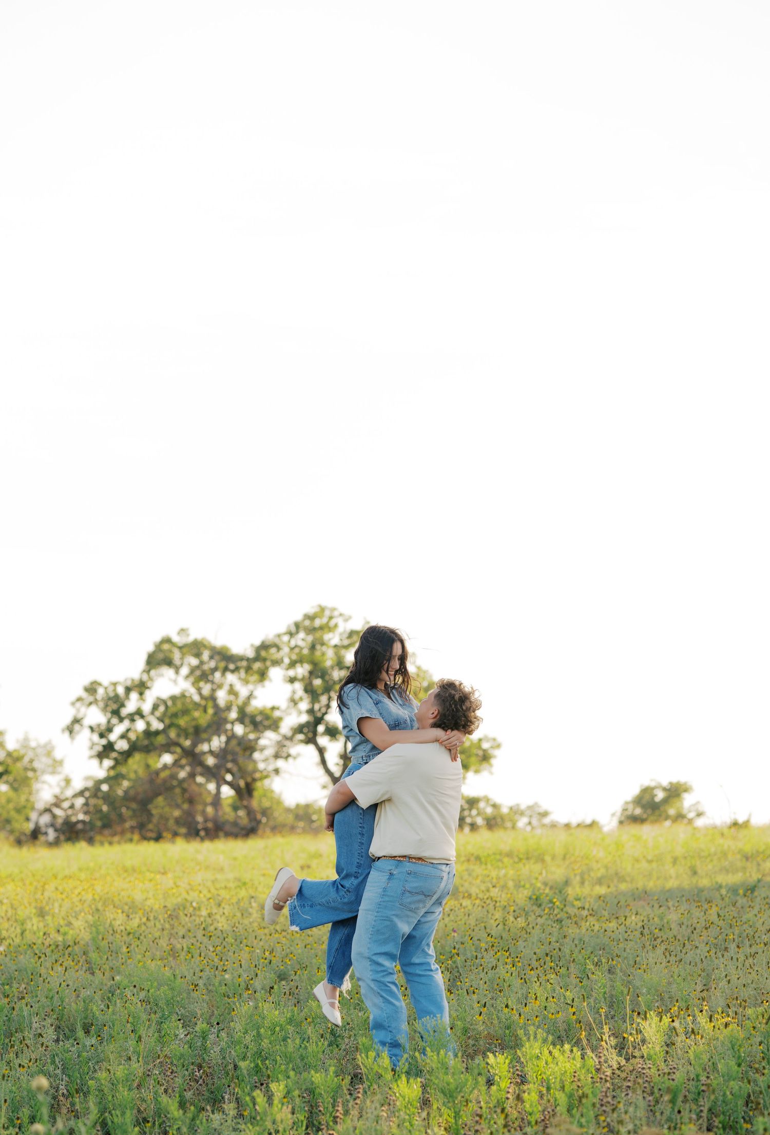 A couple embraces in a sunny green meadow, the man lifting the woman joyfully with trees in the background.