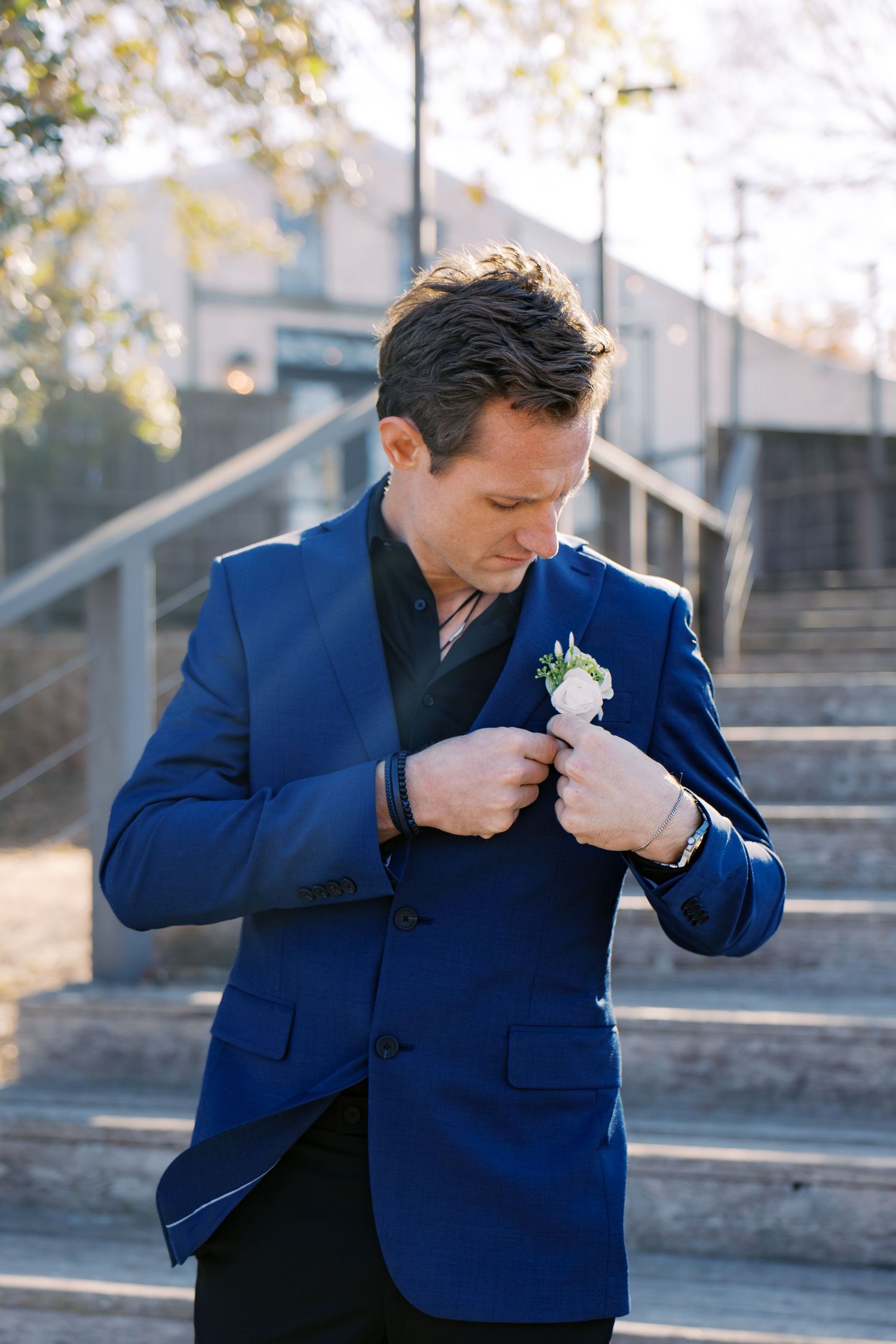 A groom in a suit adjusts his white boutonniere while standing on outdoor steps before his wedding ceremony. Oklahoma wedding photograper.
