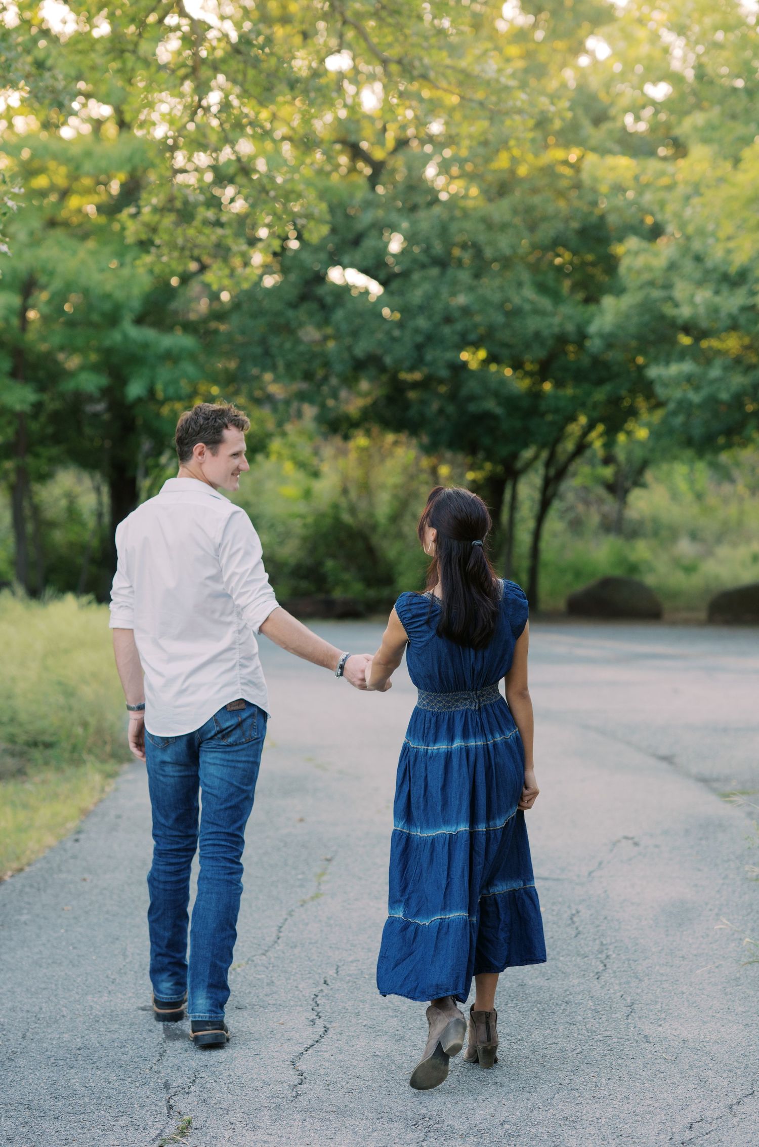 Couple holding hands walking down a tree-lined path, woman in blue maxi dress and man in white shirt and jeans.