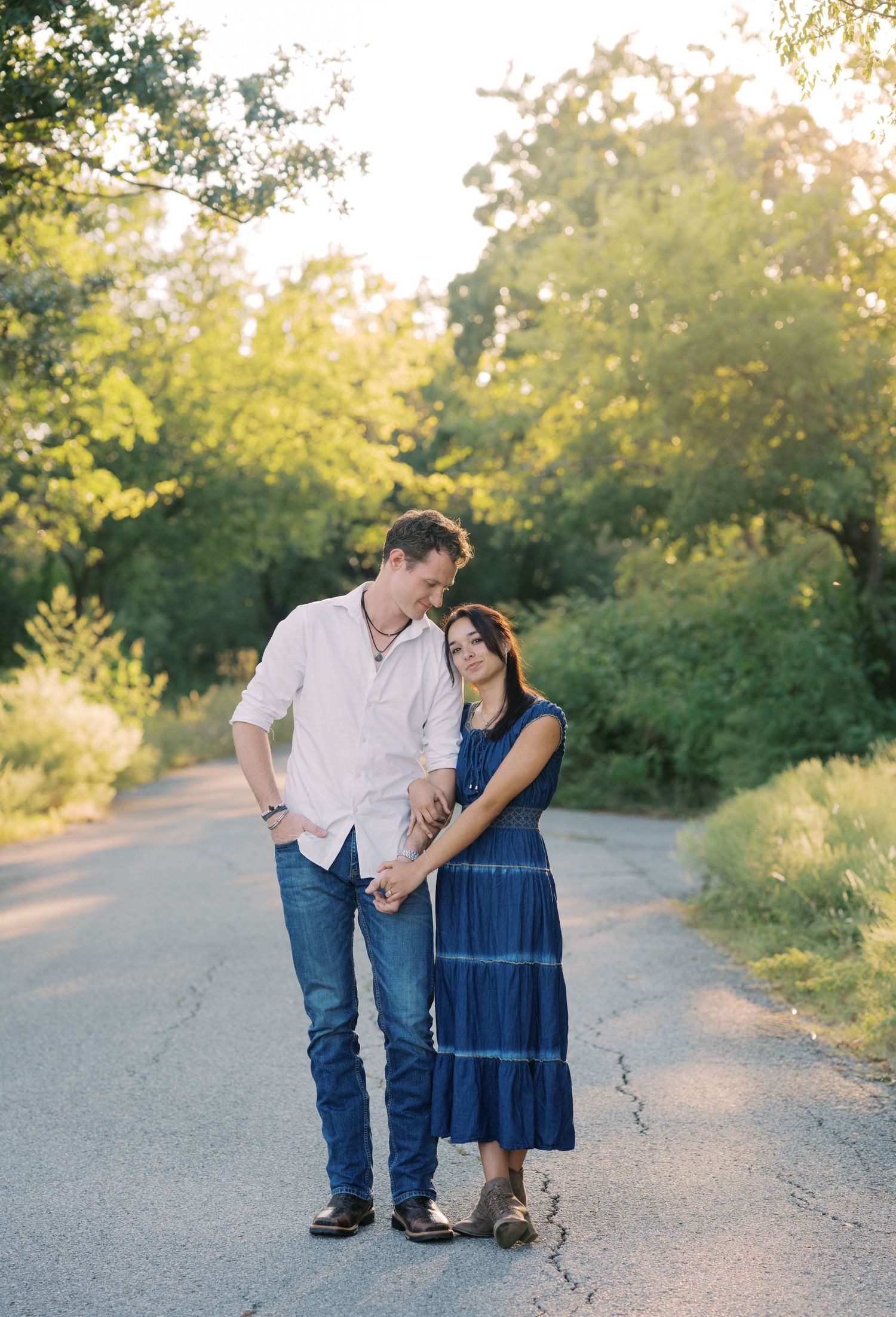 A couple stands on a sunlit tree-lined path, holding hands and gazing at each other in a romantic outdoor setting.