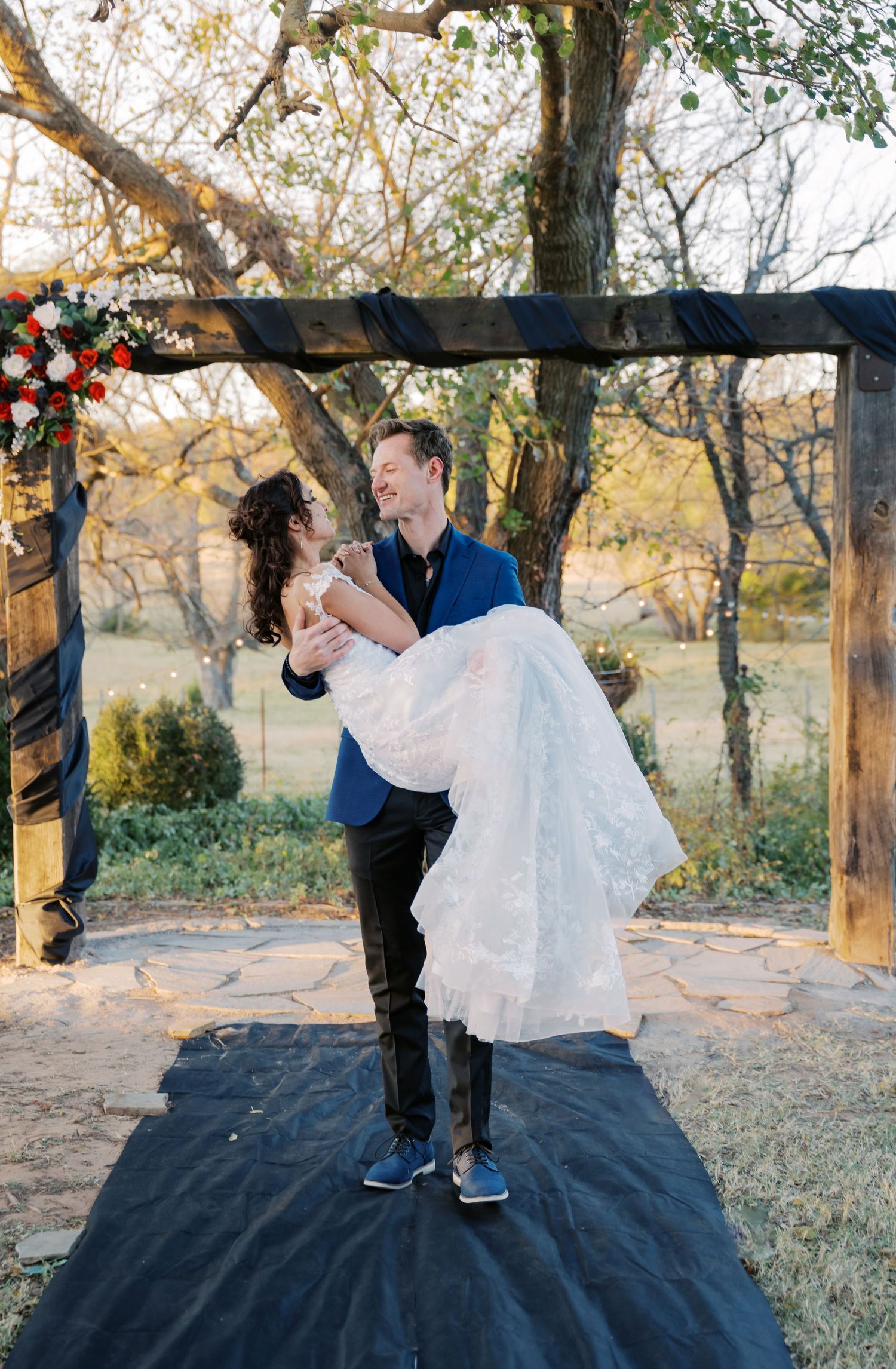 Groom in blue suit lifts bride in white lace dress under rustic wooden arch outdoors at golden hour wedding ceremony. Oklahoma wedding.