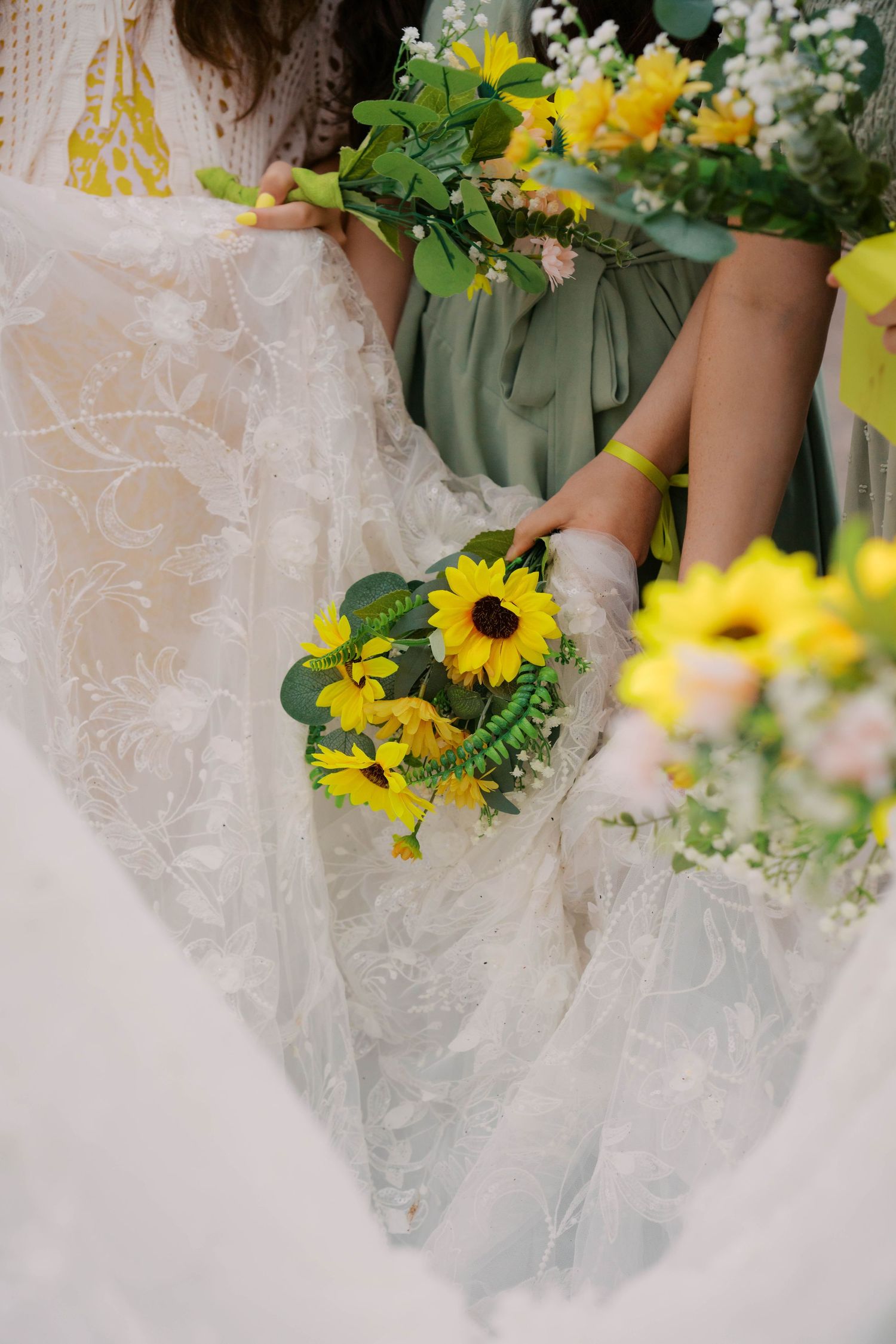 A bride in lace gown holds a sunflower bouquet beside bridesmaids in sage green dresses.