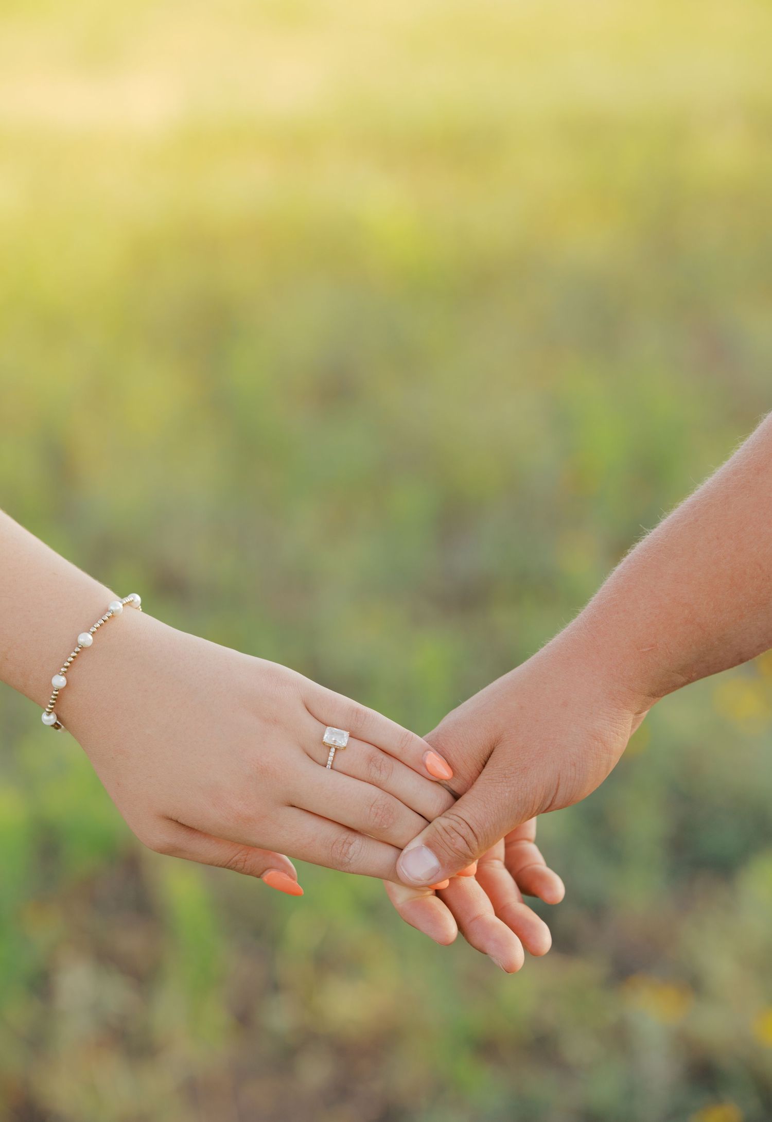 Close-up of couple holding hands displaying a diamond engagement ring and pearl bracelet outdoors.