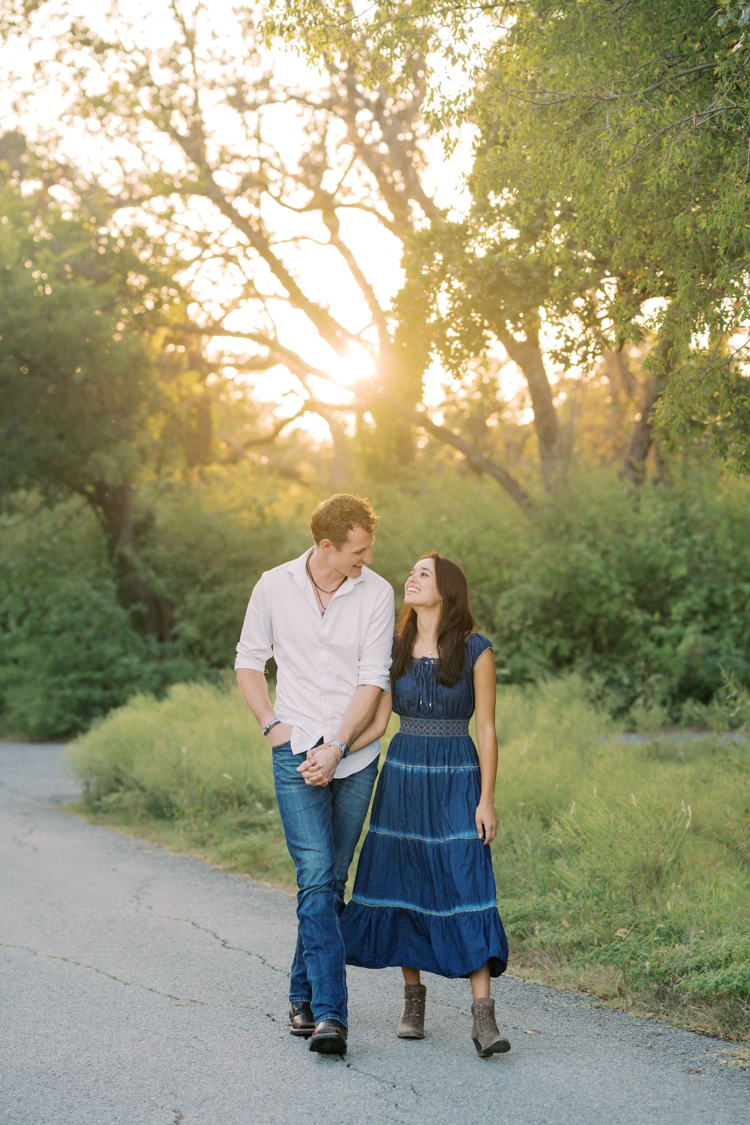 Couple walks hand in hand along a sunlit path surrounded by lush green trees at golden hour.