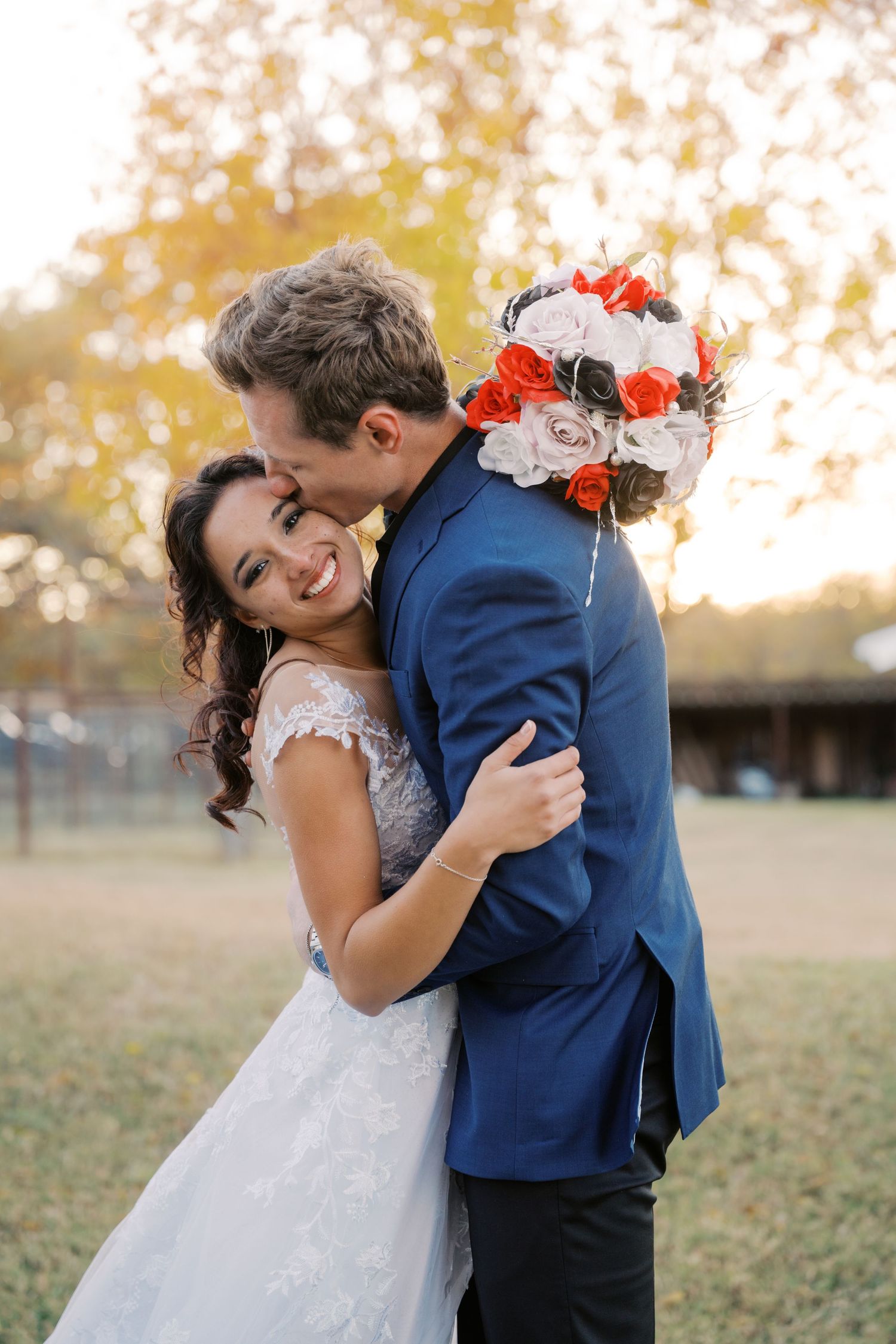 Smiling bride in lace gown embraced by groom in blue suit holding red and white rose bouquet outdoors.