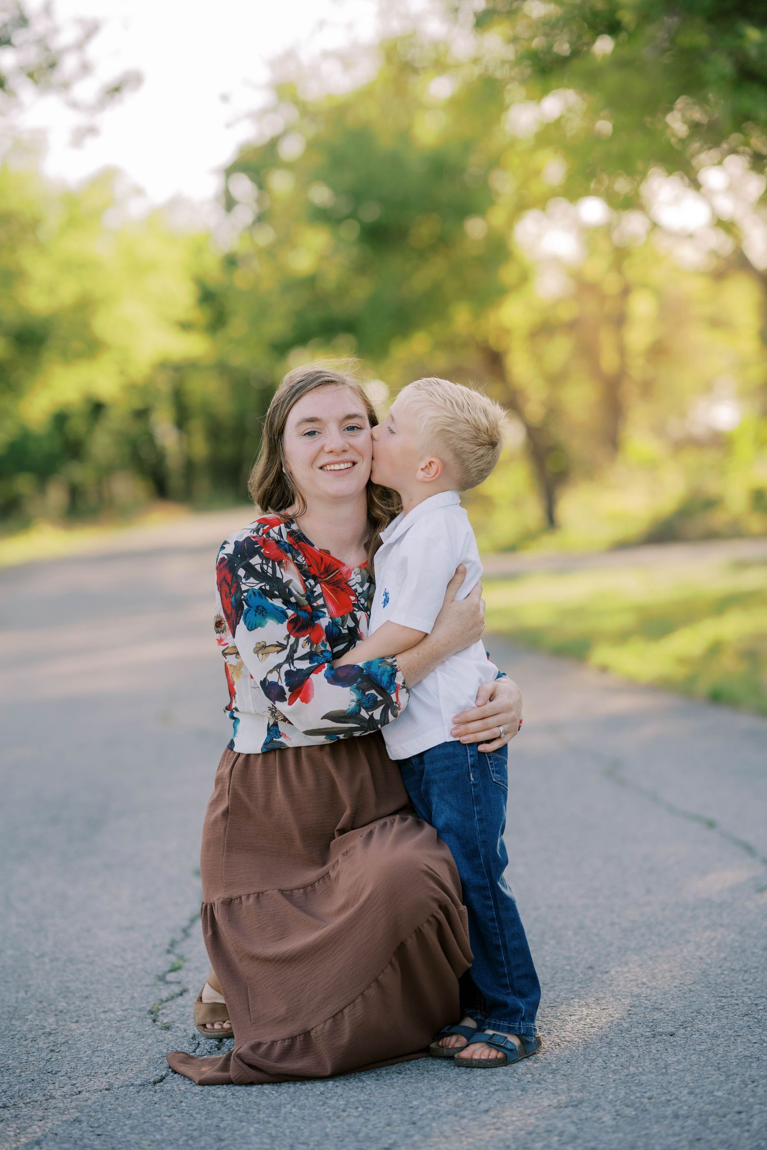 A smiling woman kneels on a road as a young blond boy hugs and kisses her cheek outdoors.